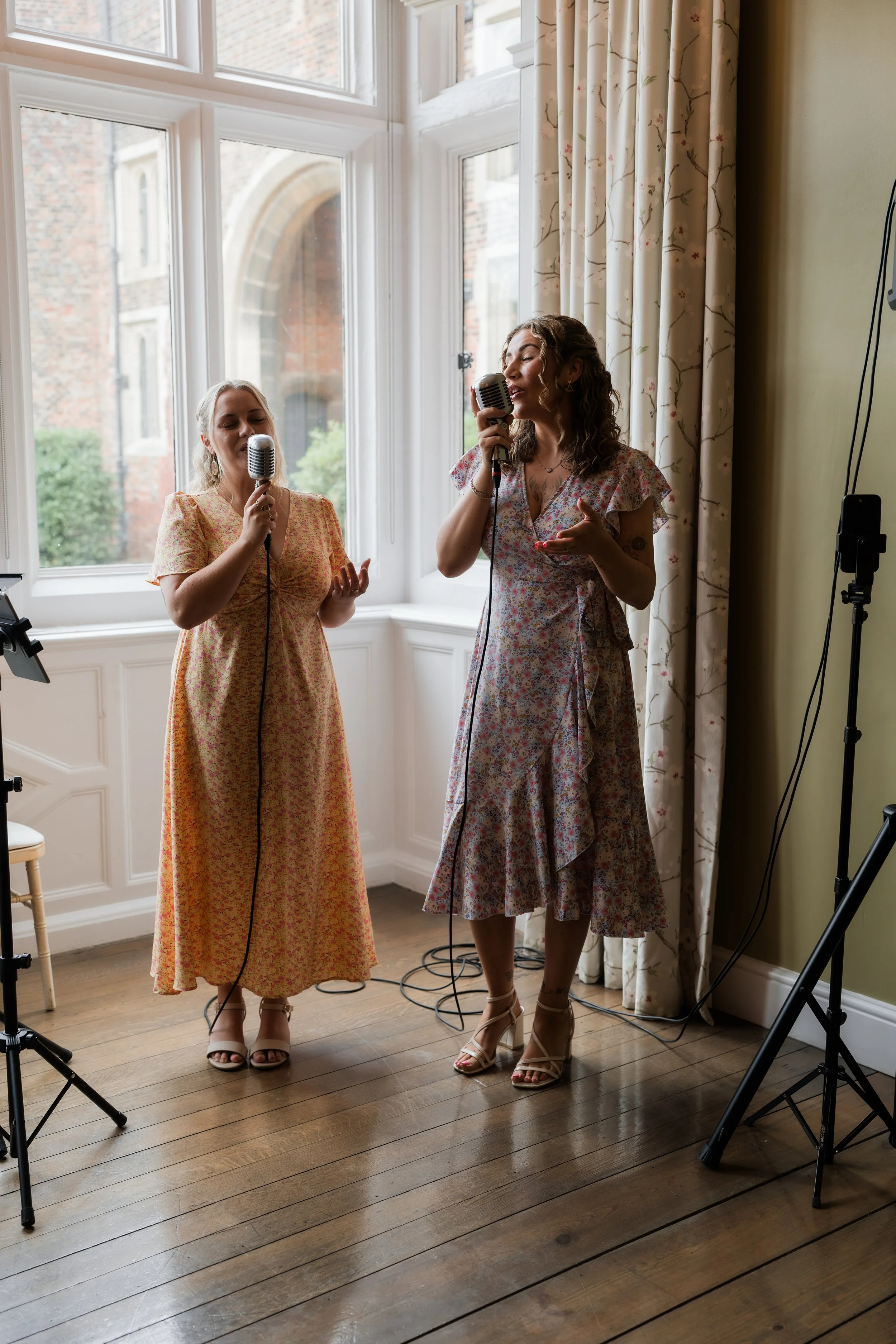 Two women singing into vintage-style microphones, standing near a large window with natural light coming in, in a cozy, well-lit interior with hardwood floors and floral curtains.