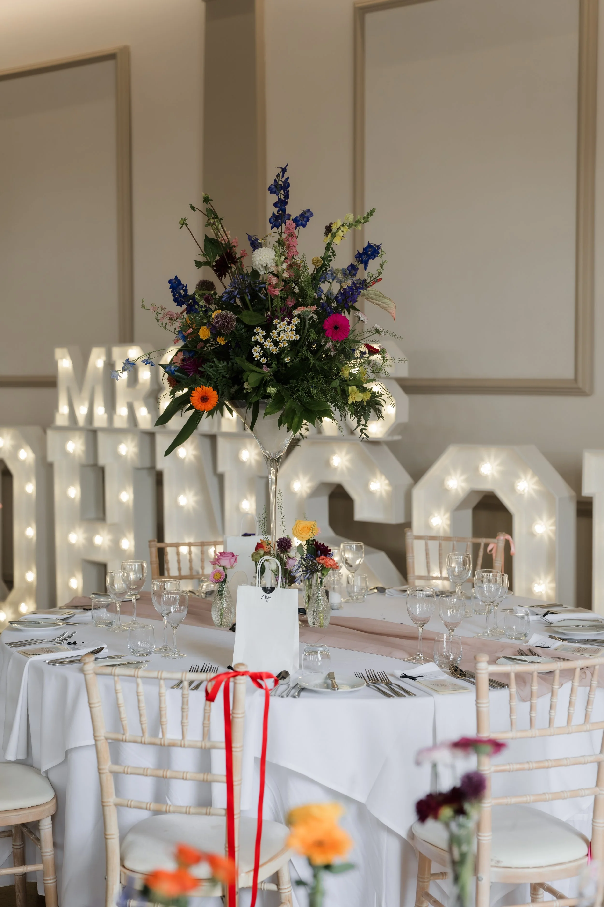 Elegant table setup for a celebration featuring a large centerpiece of colorful flowers in a tall glass vase, surrounded by wine glasses, plates, and cutlery at a decorated event space with large illuminated letters in the background.