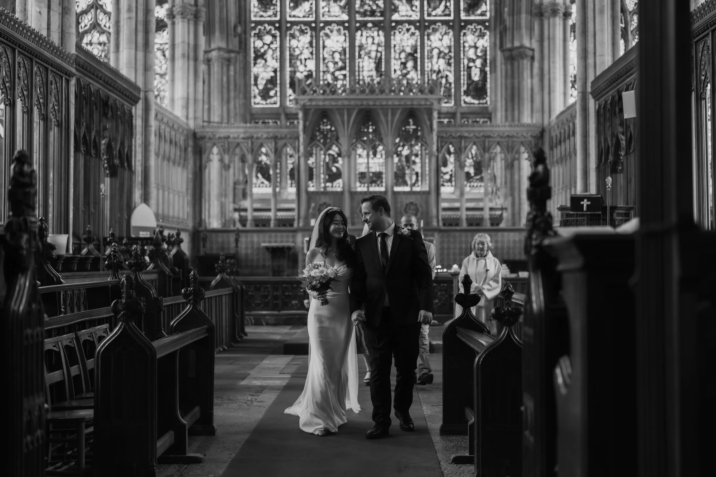 A bride and groom walk down the aisle inside Hull Minster, smiling at each other, with guests in the background.