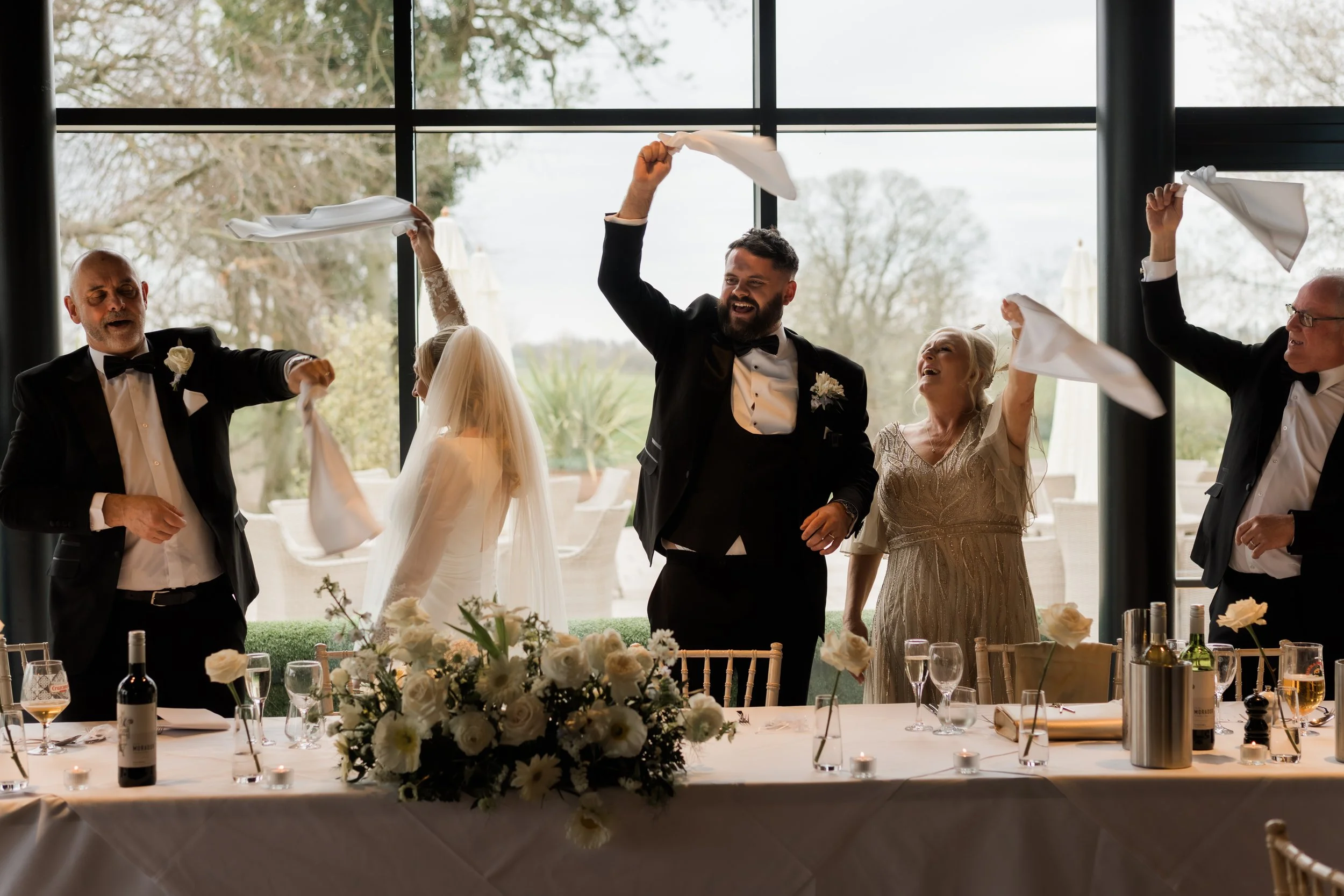 Wedding celebration with people dancing, a bride in a wedding dress and veil, and a groom in a tuxedo, at a decorated table with flowers and wine bottles, inside a venue with large windows and trees outside.