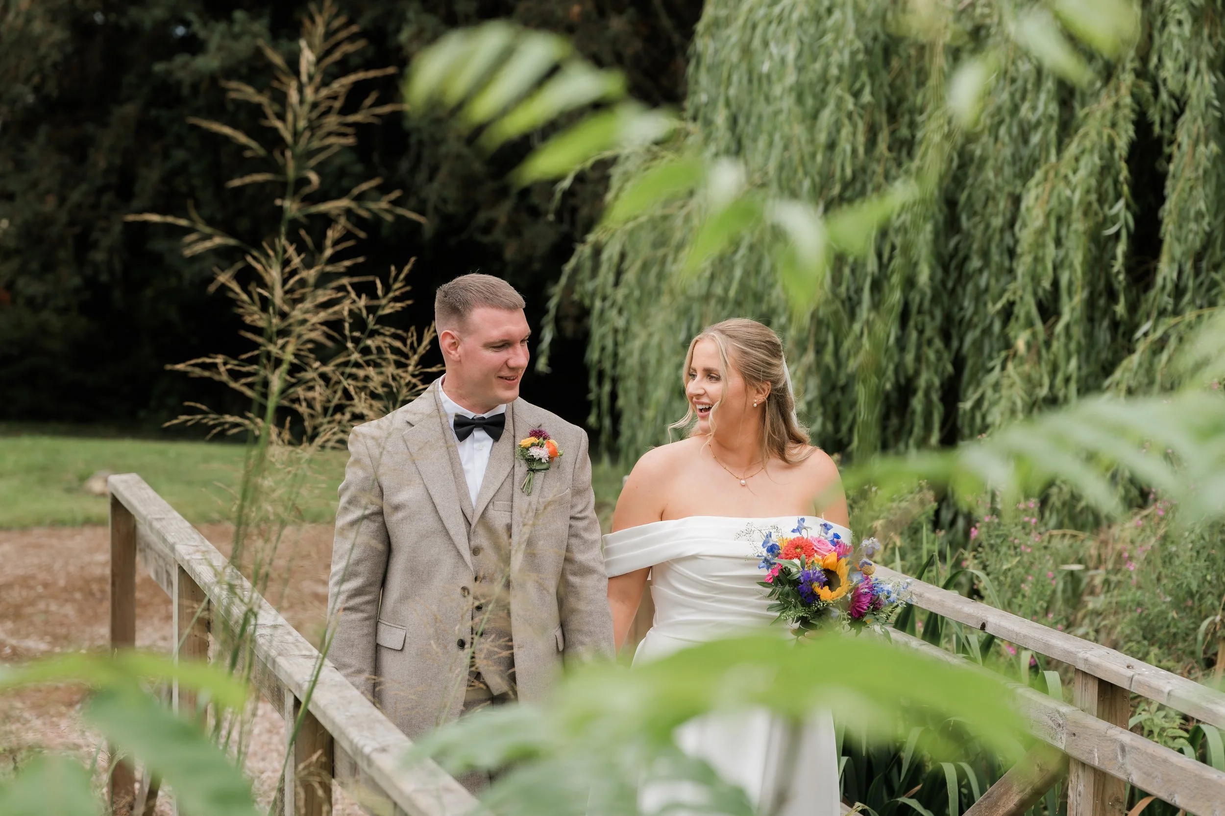 A bride and groom walking together outdoors on a wedding day, surrounded by greenery and trees, smiling at each other. The bride holds a colorful bouquet, and the groom wears a light gray suit with a black bow tie.