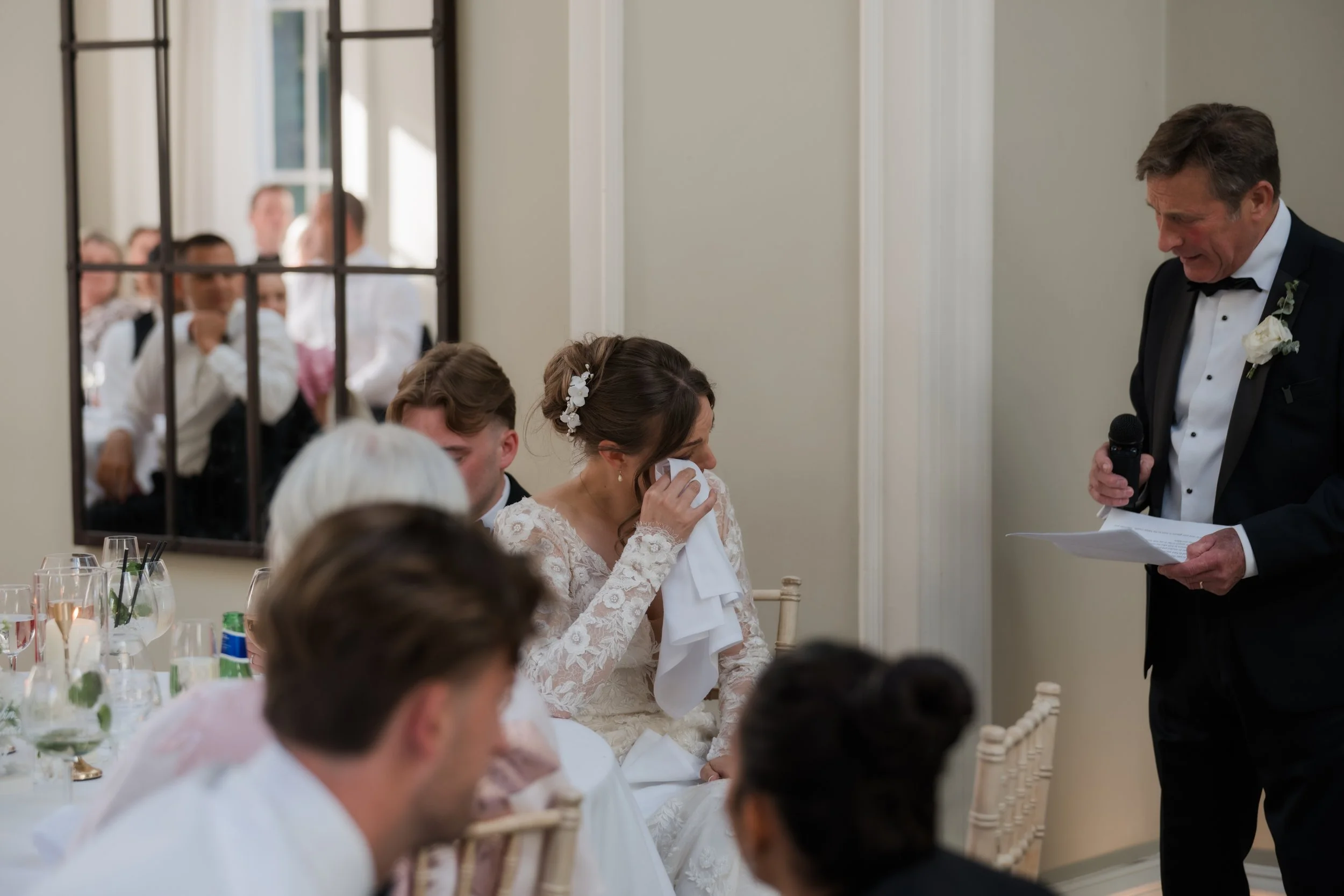 A wedding reception scene with a bride wiping tears and a groom reading vows to her. Guests are seated at a decorated table with glasses and utensils, and a man in a tuxedo is speaking into a microphone.