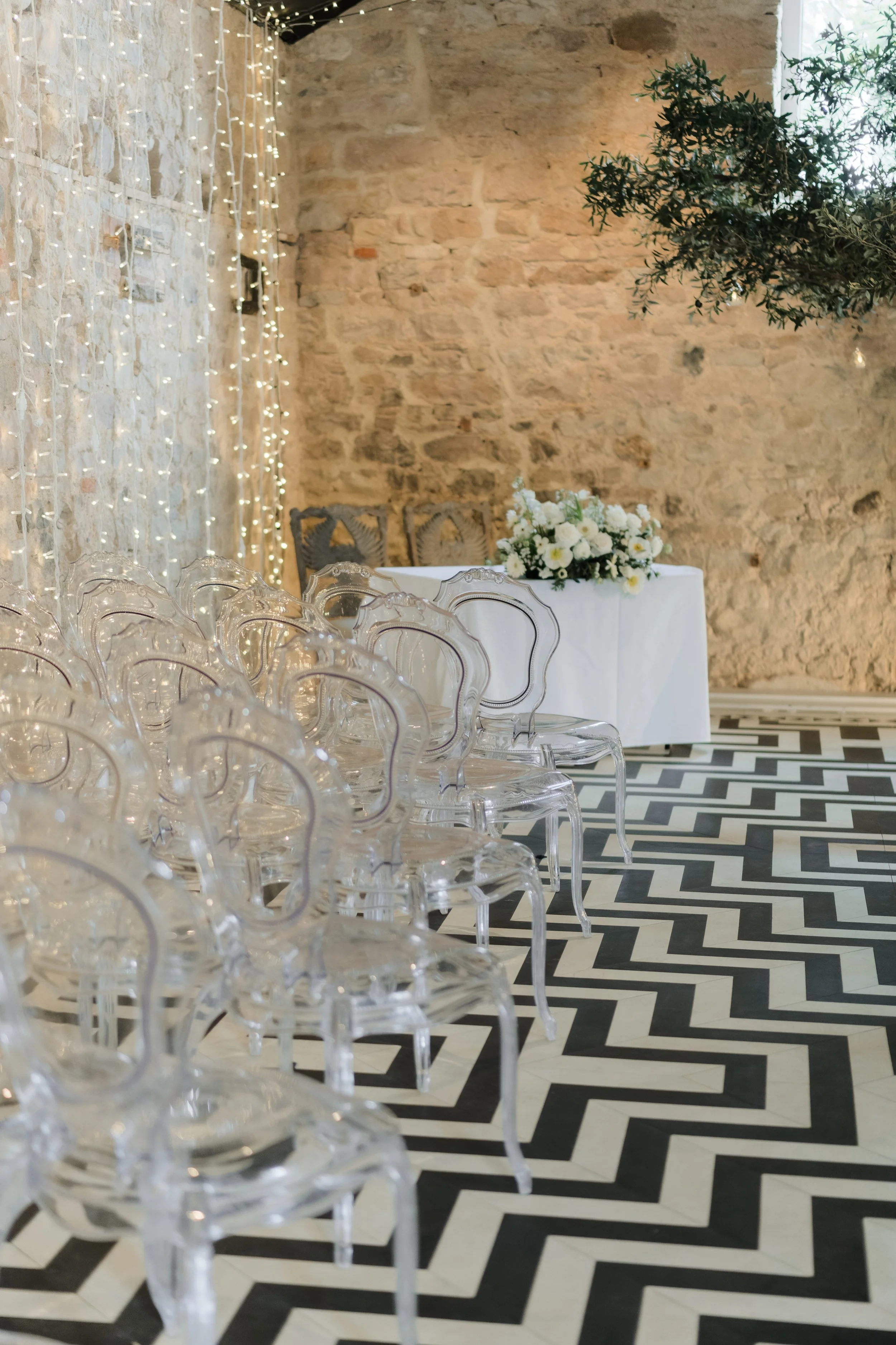 Elegant event setup with clear ghost chairs arranged in rows, a black and white geometric patterned floor, a table with white cloth and floral centerpiece, and string lights hanging against a rustic brick wall.
