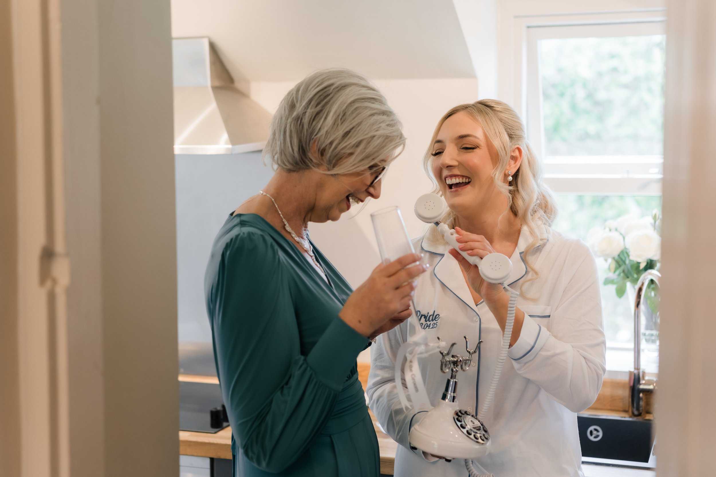 A joyful bride in a robe talking on a vintage white rotary phone held by a smiling woman in a teal dress, in a well-lit kitchen with a window and white roses in the background.