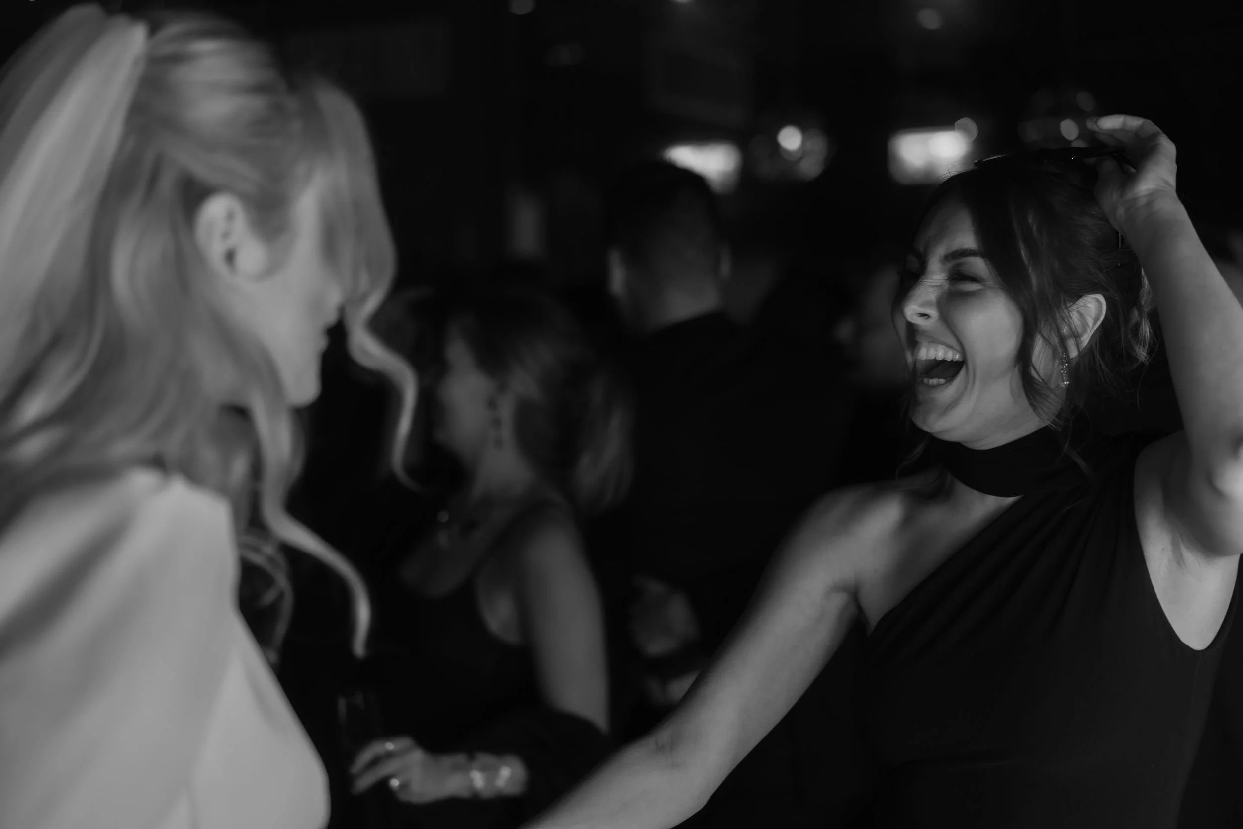 Two women laughing and enjoying themselves at a social gathering. One woman has blonde hair and the other has dark hair and is wearing a black dress.