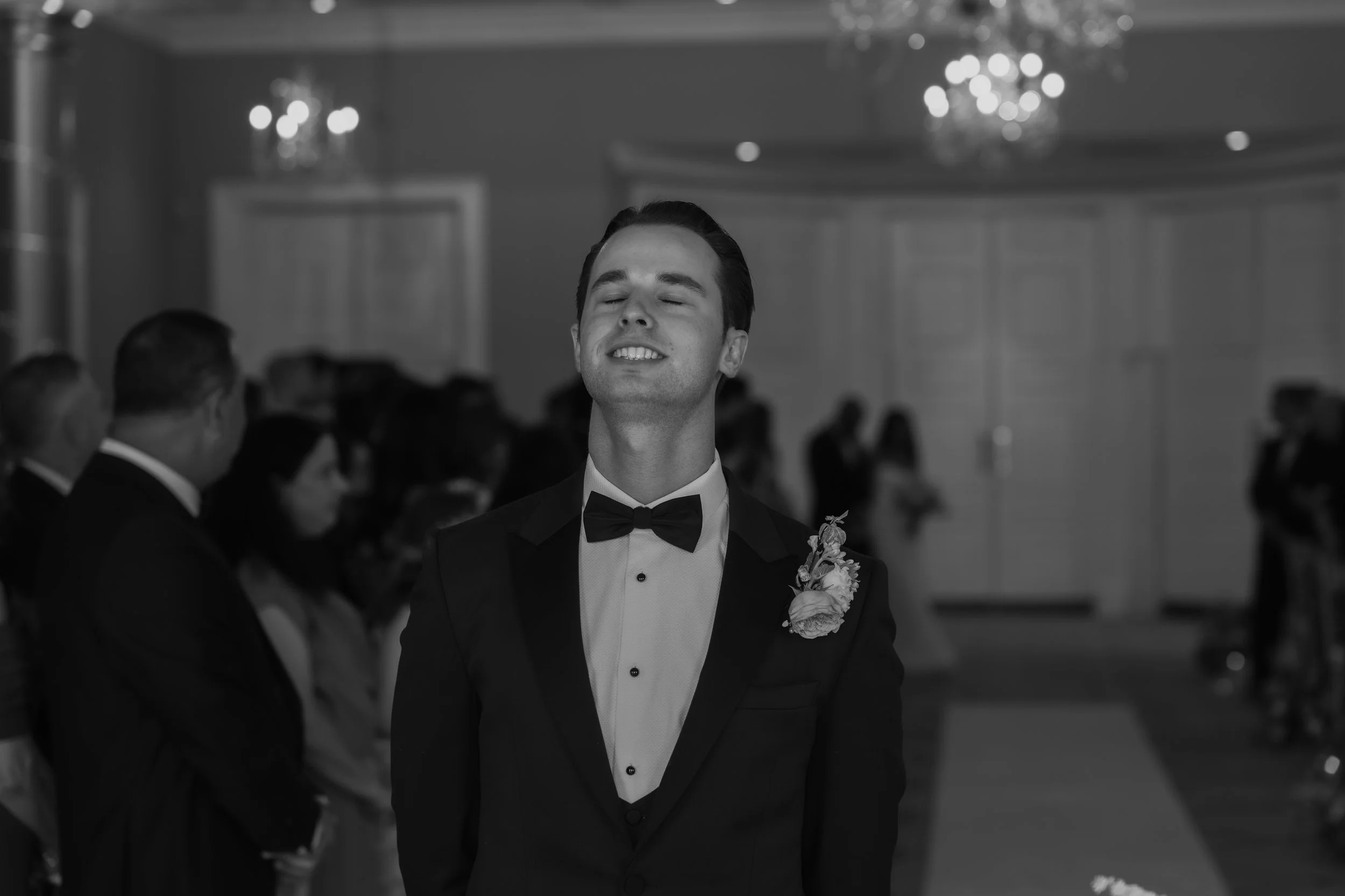 A man in a tuxedo with a bow tie and a boutonniere standing with his eyes closed at a formal event, with other guests seated in the background.