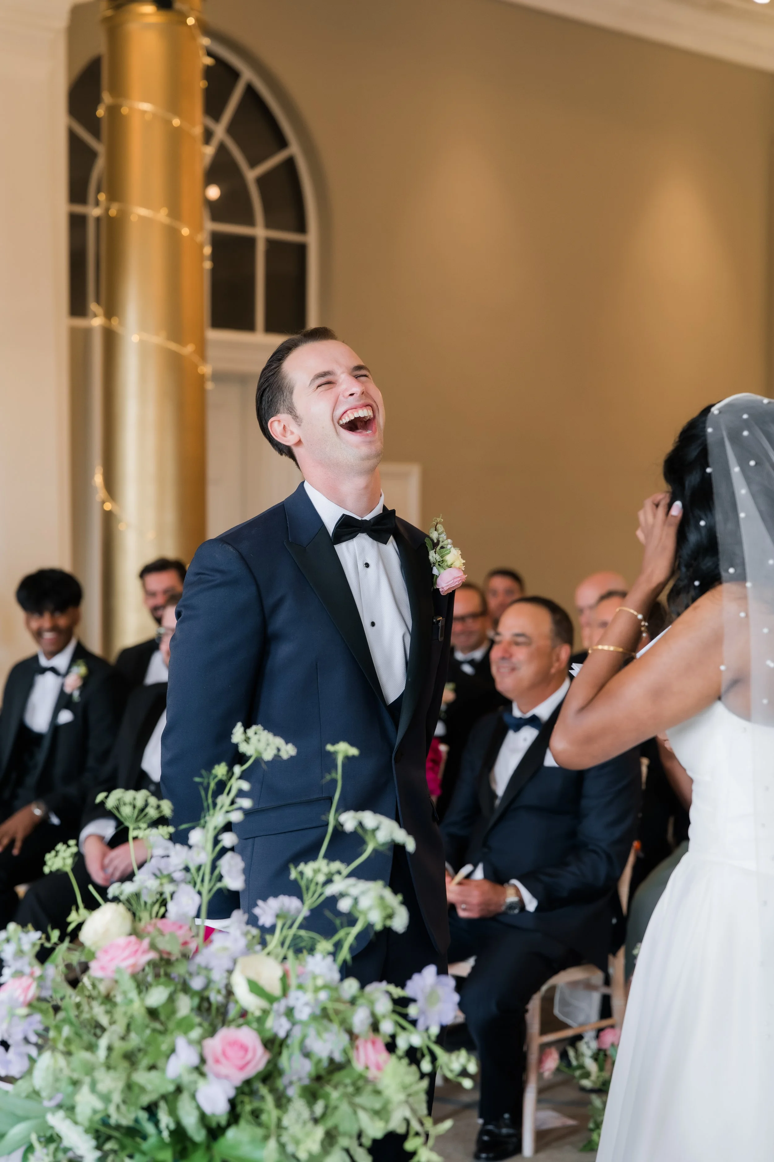 A groom in a black tuxedo and bow tie is laughing during a wedding ceremony, surrounded by seated guests in formal attire, with a floral arrangement in the foreground and a gold column and arched window in the background.