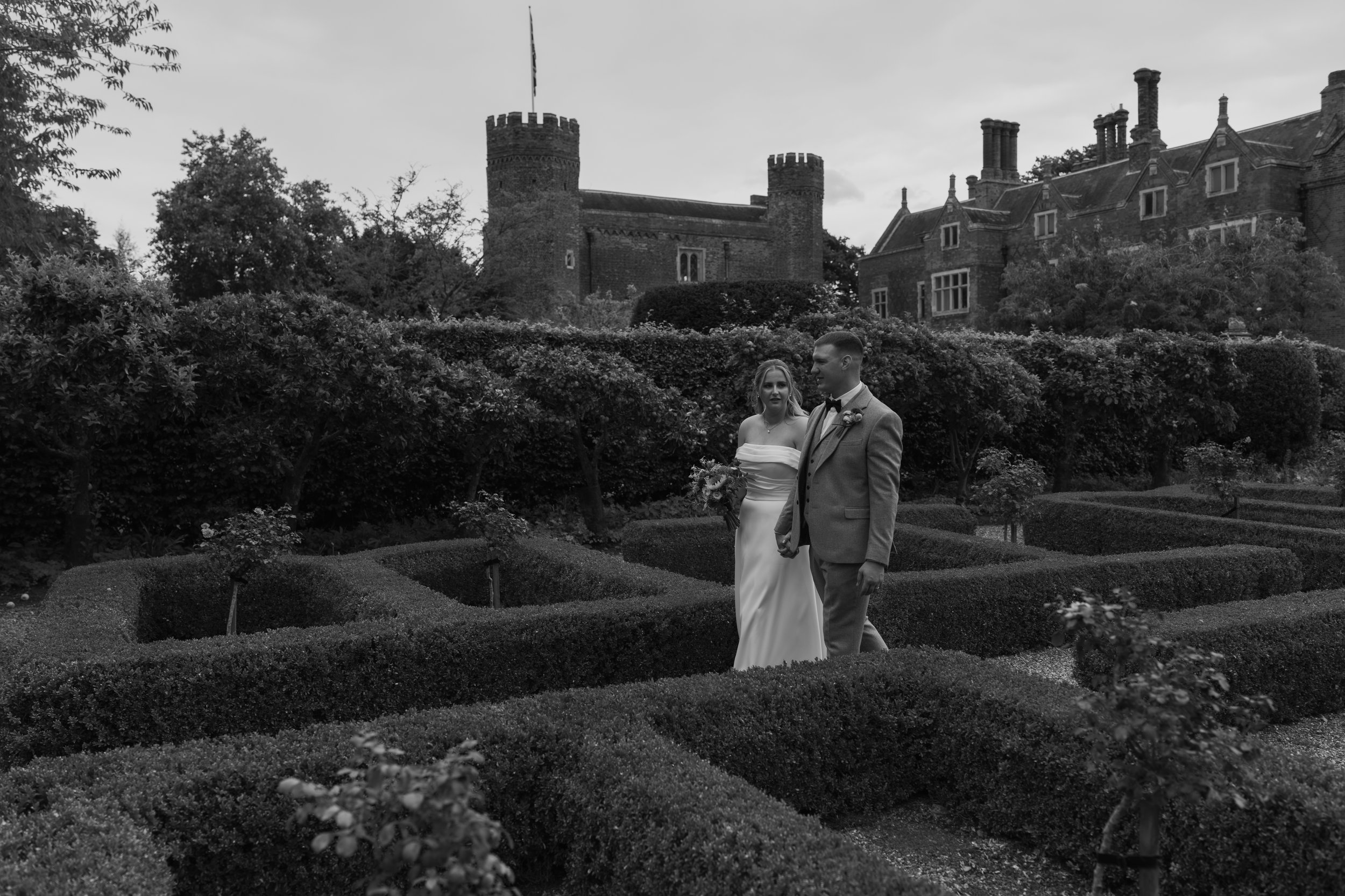 A black and white photo of a bride and groom holding hands in a garden with neatly trimmed hedges and a castle in the background.