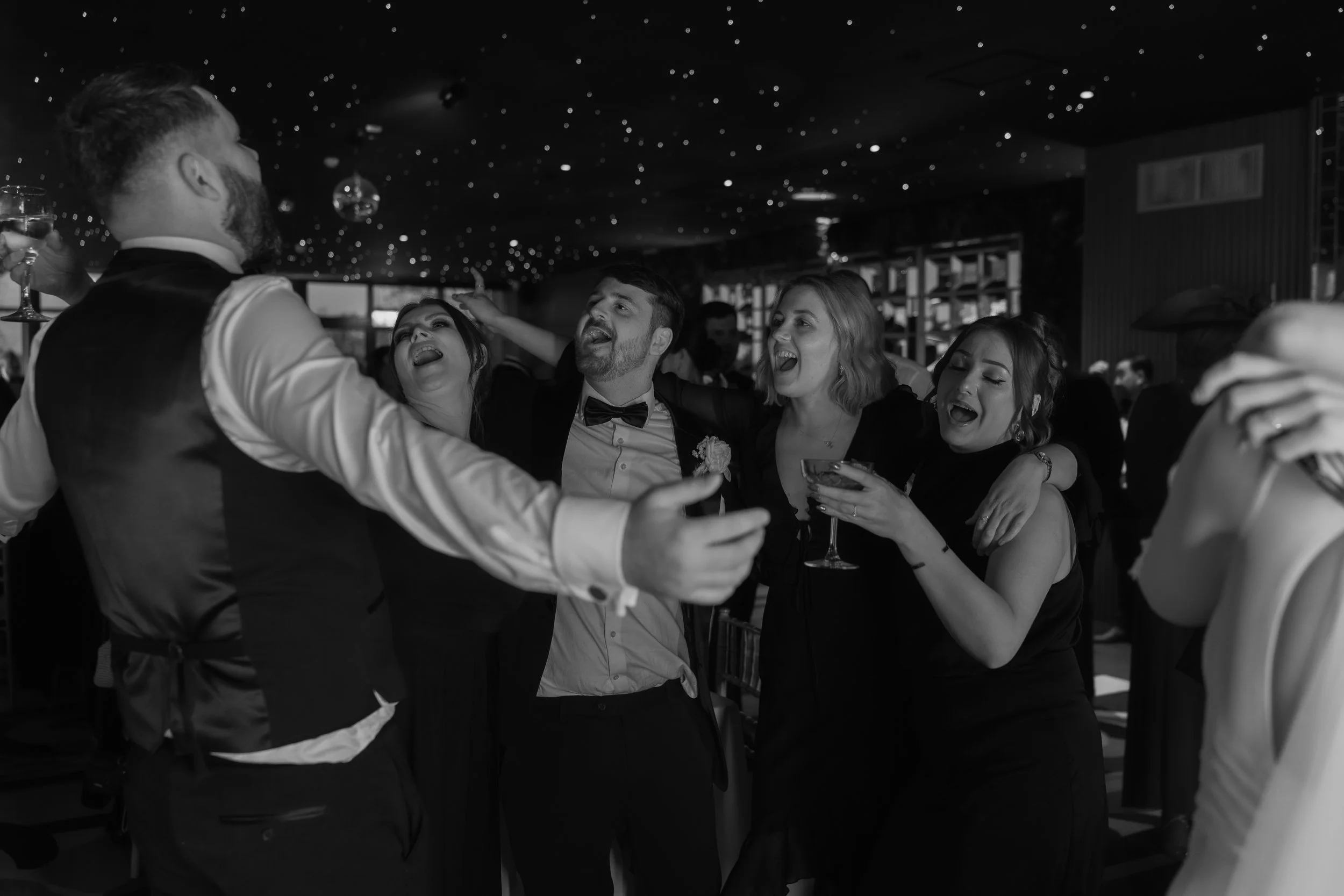 A group of people celebrating at a wedding reception, dancing and enjoying drinks in a decorated indoor venue.