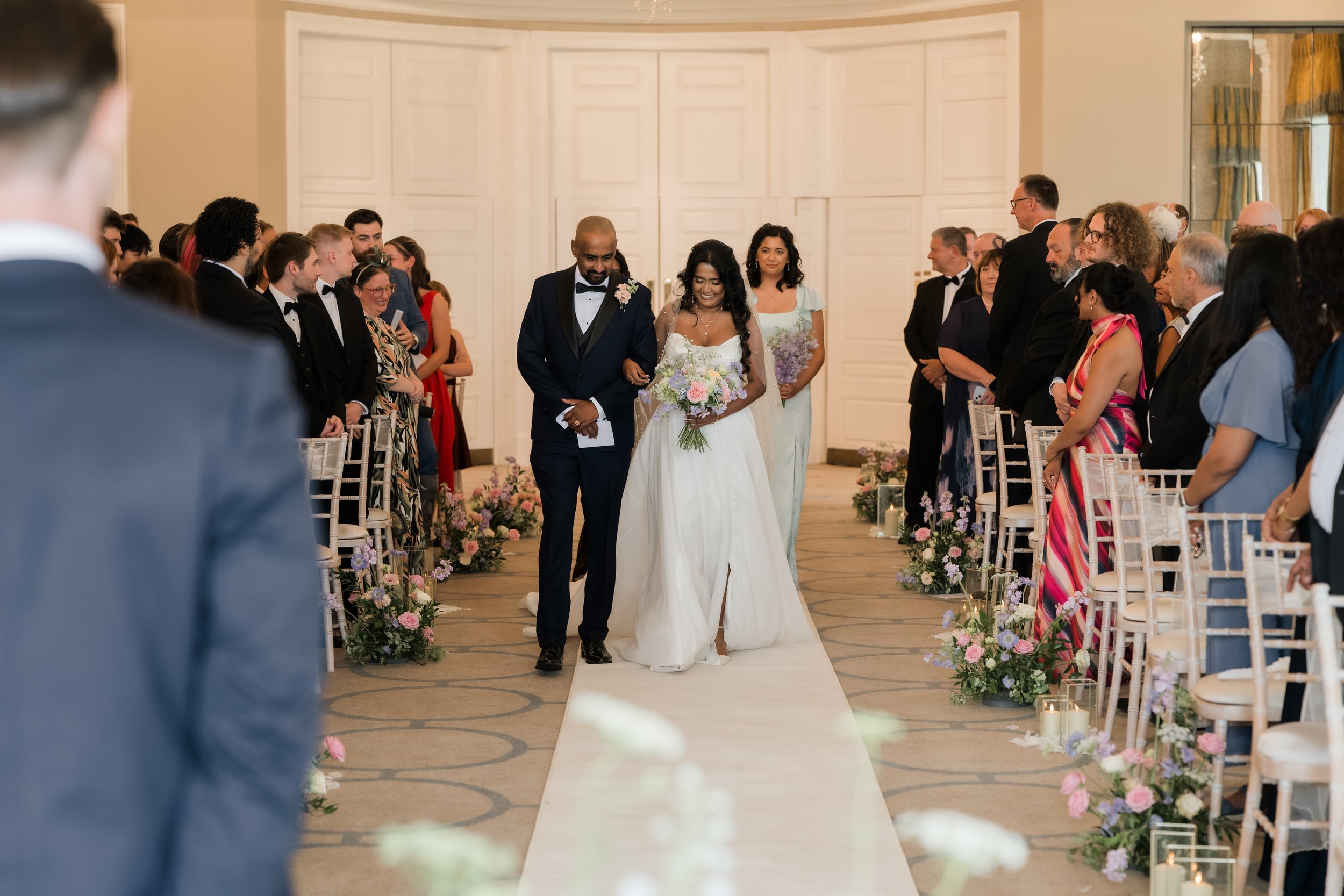 A bride walking down the aisle assisted by a man, possibly her father, at a wedding ceremony with guests seated on both sides, floral arrangements, and candles.