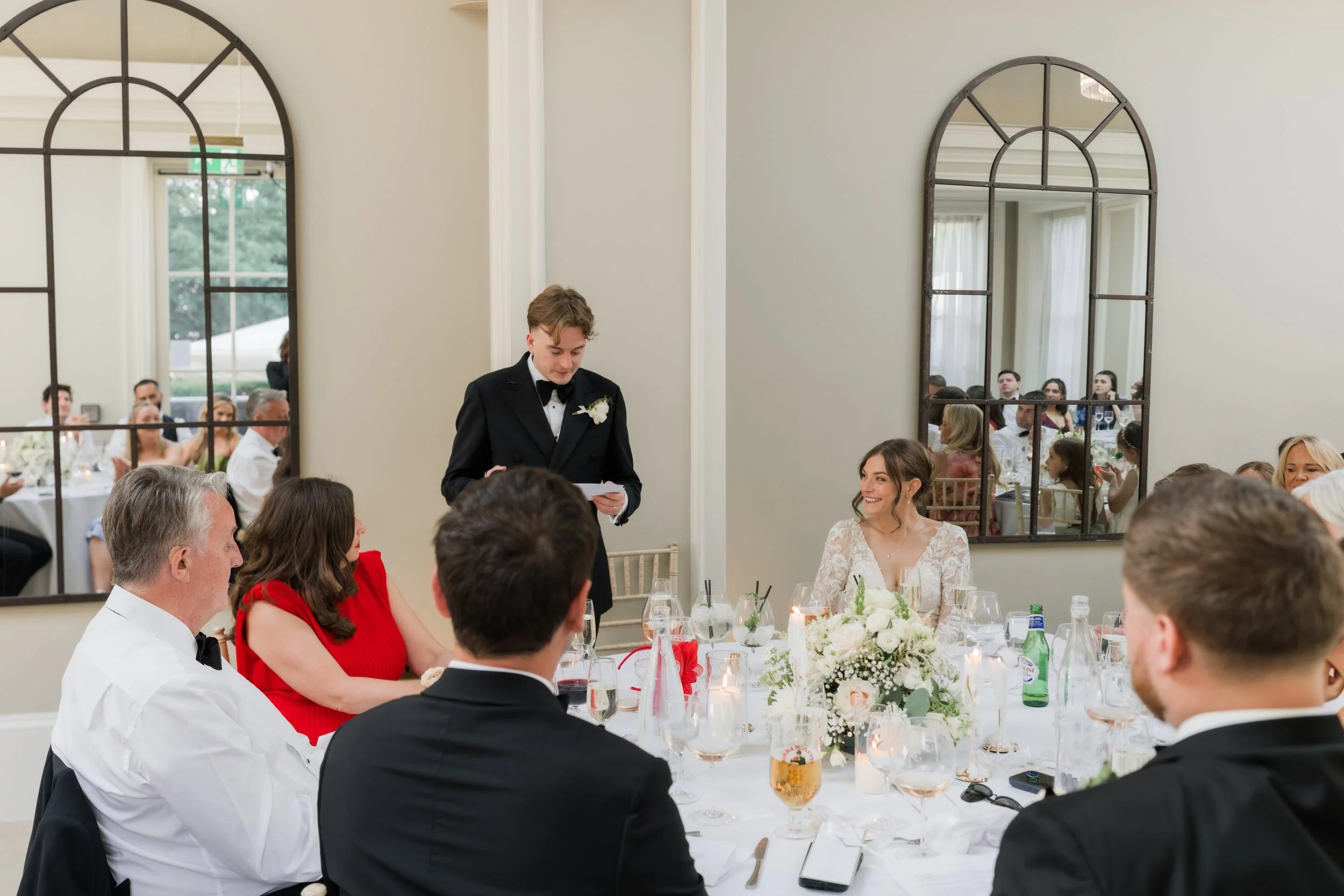 A wedding reception with guests seated around a decorated table, a man in a tuxedo giving a speech, and a smiling bride with long hair in a lace wedding dress. Mirrors on the wall reflect more guests.