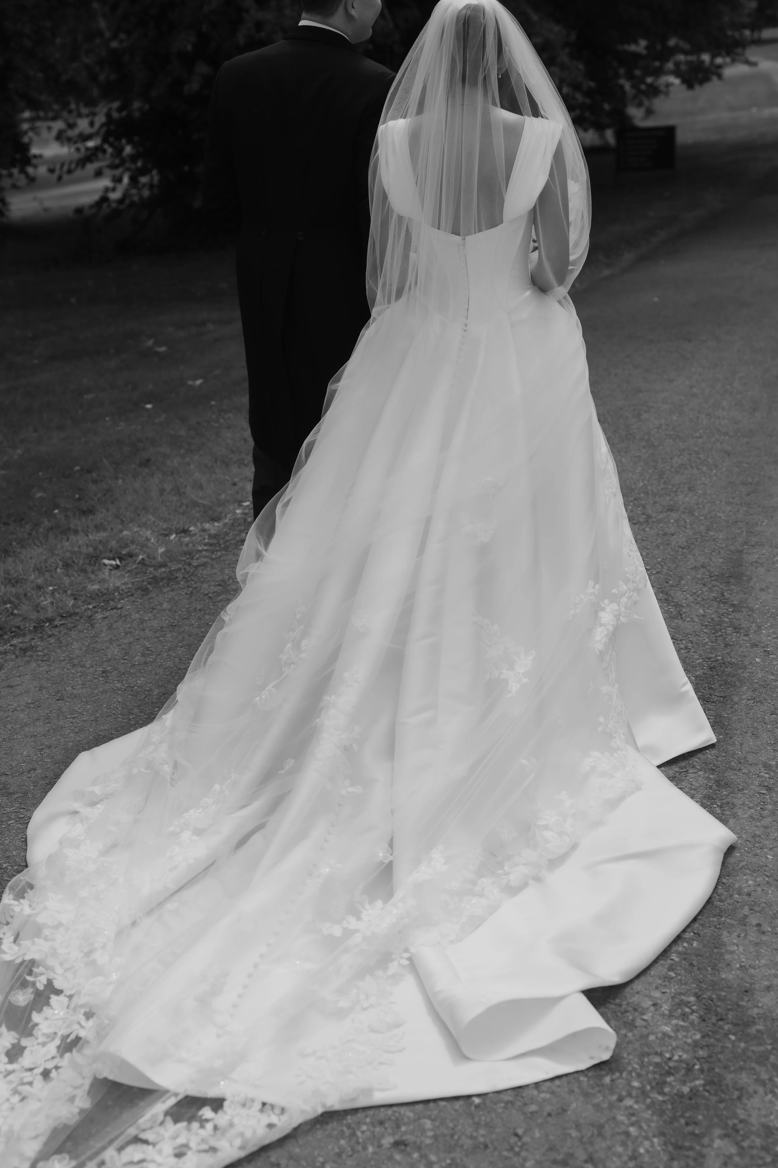 Black and white photo of a bride and groom standing outdoors facing away, with the bride wearing a long wedding gown with a train and veil, and the groom in a suit, surrounded by trees and grass.