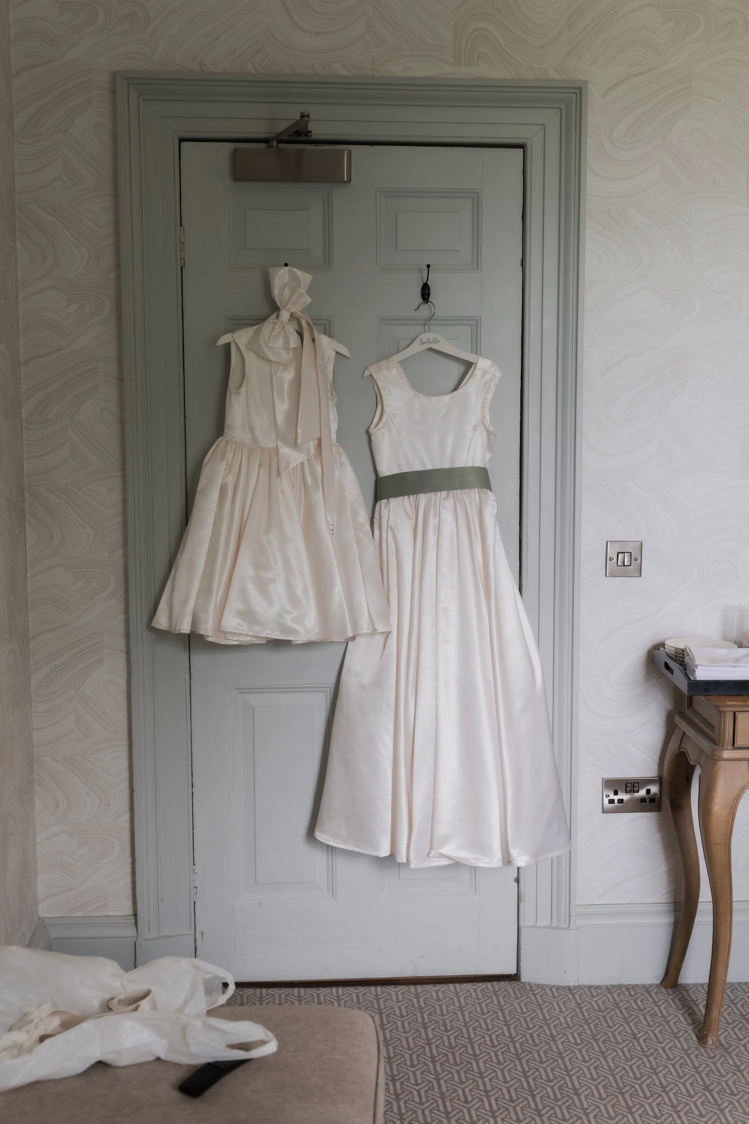 Two white dresses hanging on a door, one with a bow and the other with a grey sash, likely for a wedding or special occasion, with some items on a nearby table.