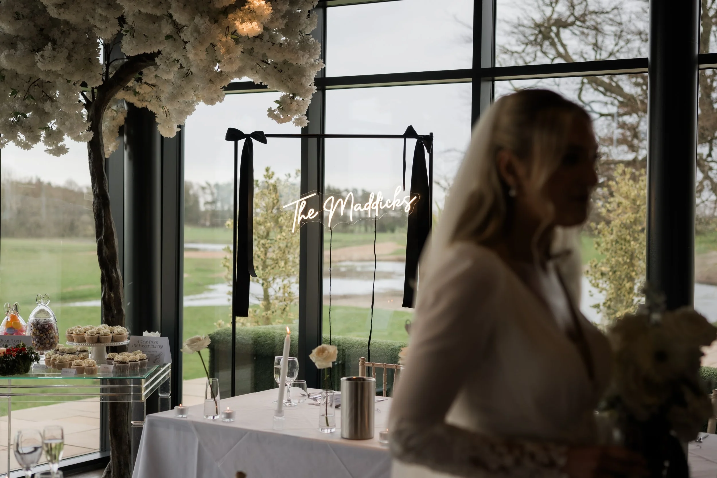 Silhouette of a woman holding a bouquet, standing indoors near decorated tables with desserts and candles, in front of large windows overlooking a green outdoor landscape, with a neon sign reading 'The Maddicks' in the background.
