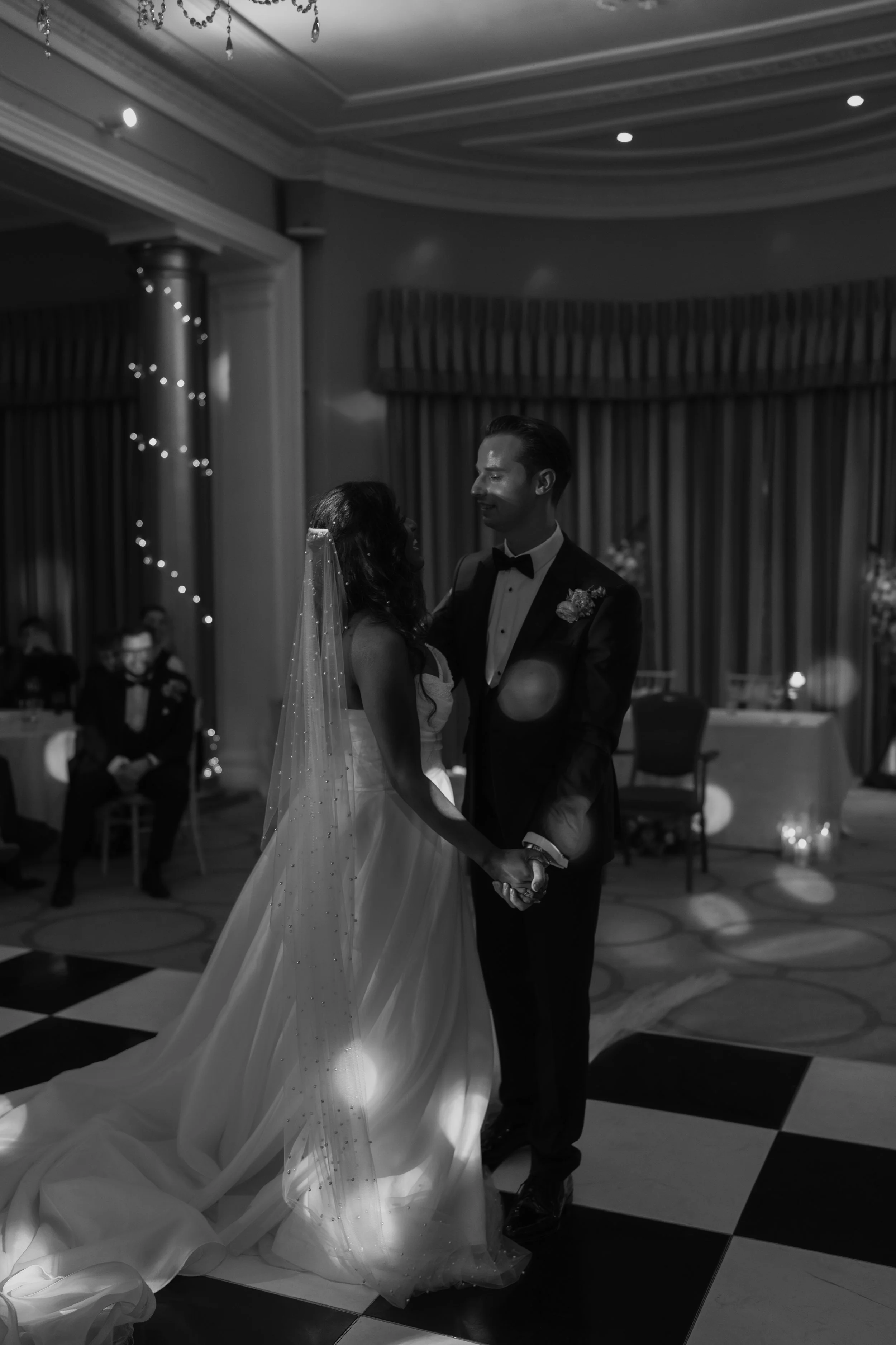 A black and white photo of a bride and groom dancing at their wedding reception, holding hands and smiling at each other.