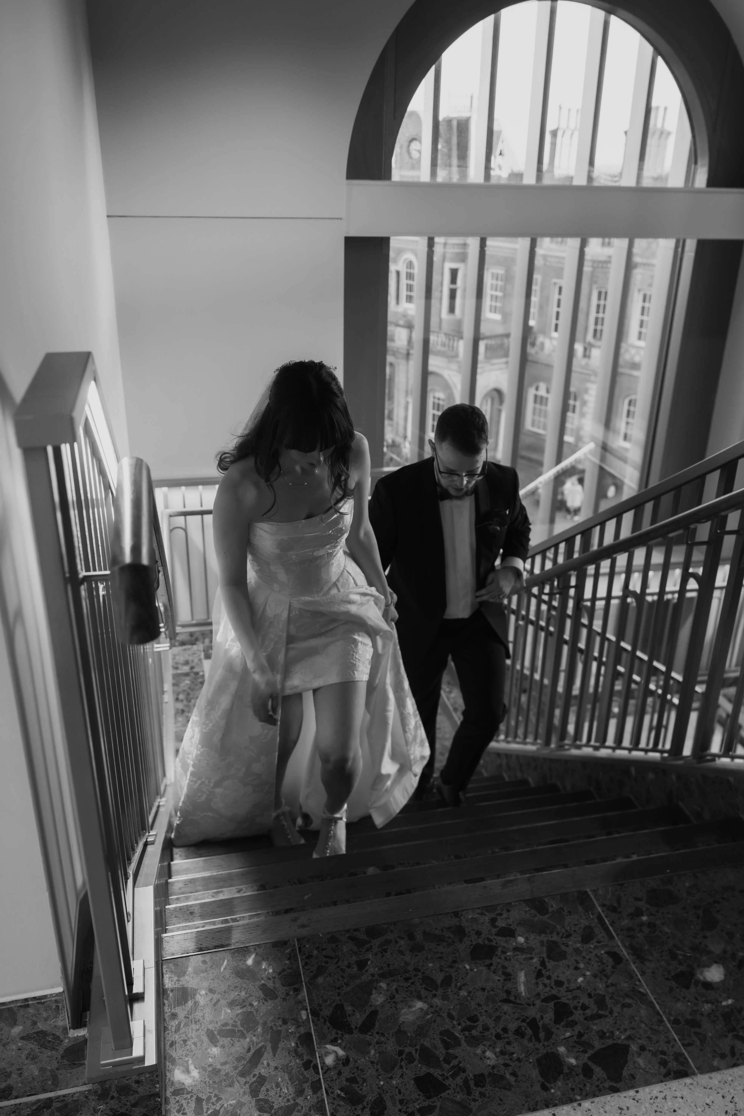 A bride and groom are ascending a staircase inside a building, with the bride lifting her dress slightly as they walk up the stairs. The scene is in black and white, with large windows in the background showing a cityscape.