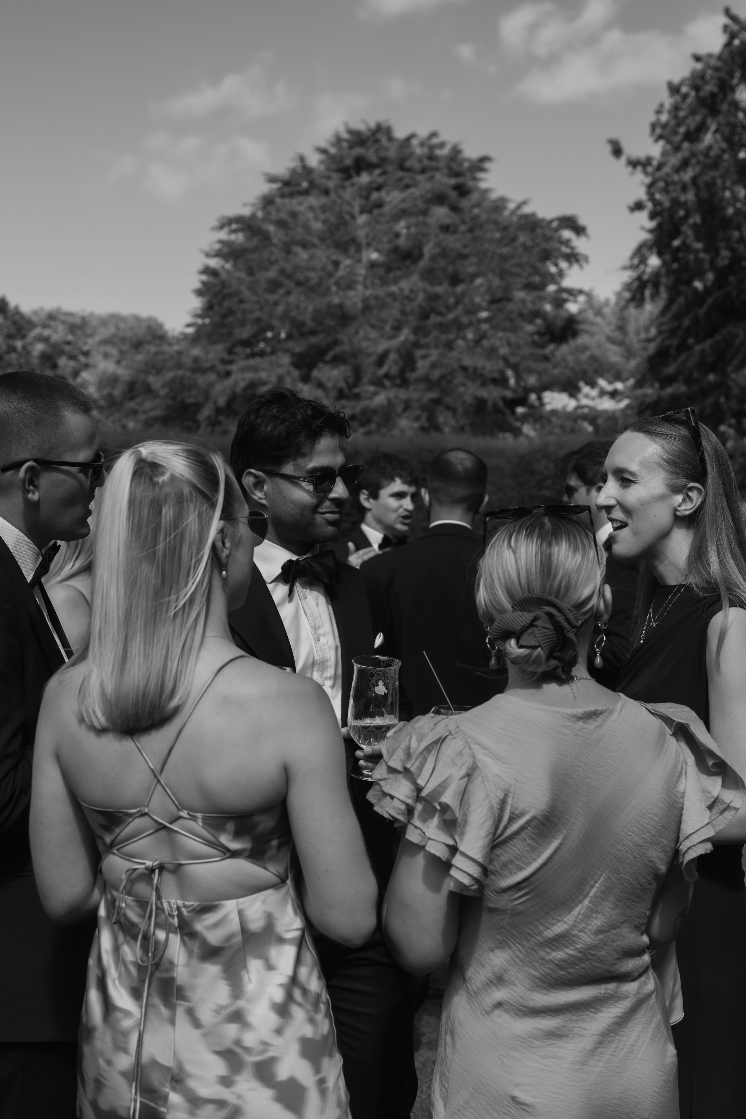 A group of people at an outdoor gathering, engaging in conversation, with trees and a sky in the background.