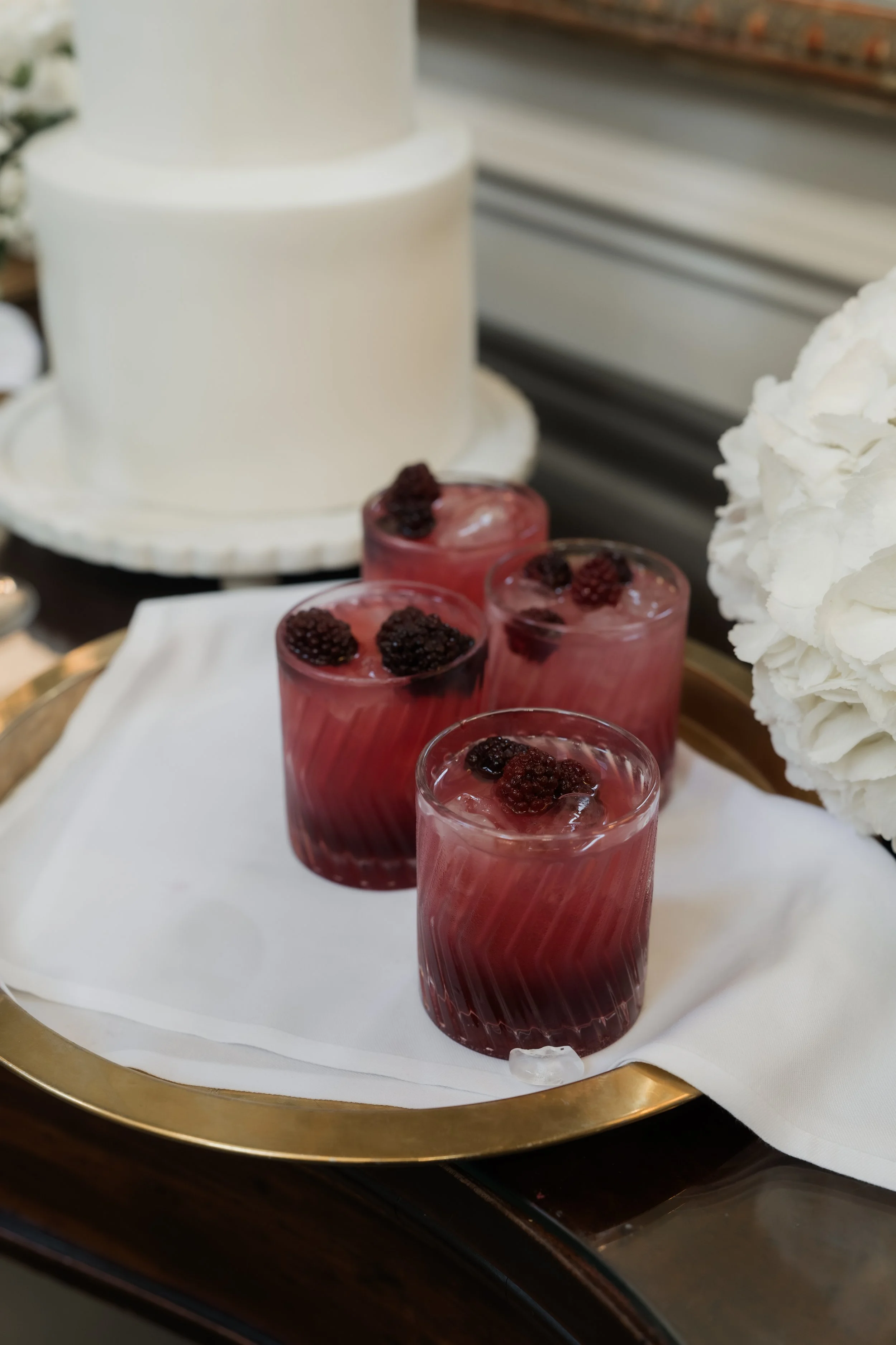 A white cake on a decorative tray and four glasses of a pink beverage with berries on top, all placed on a white tablecloth.