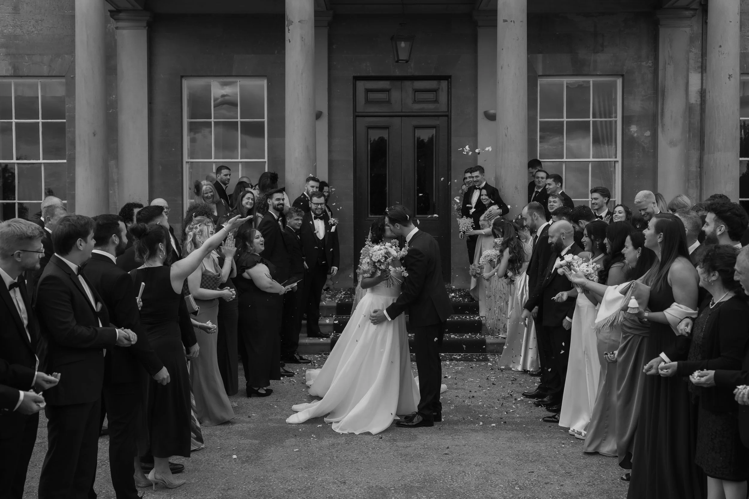 Black and white photo of a wedding celebration with a bride and groom kissing in front of a large group of guests gathered outside a historic building with columns, large windows, and a wooden door.