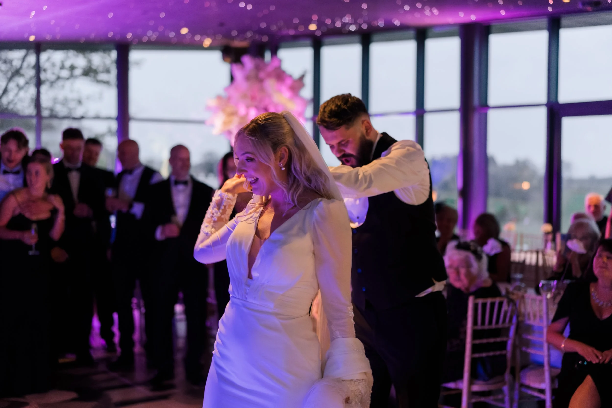 A bride and groom sharing a dance at their wedding reception, with guests watching and taking photos in the background, in a decorated indoor venue with large windows and purple lighting.