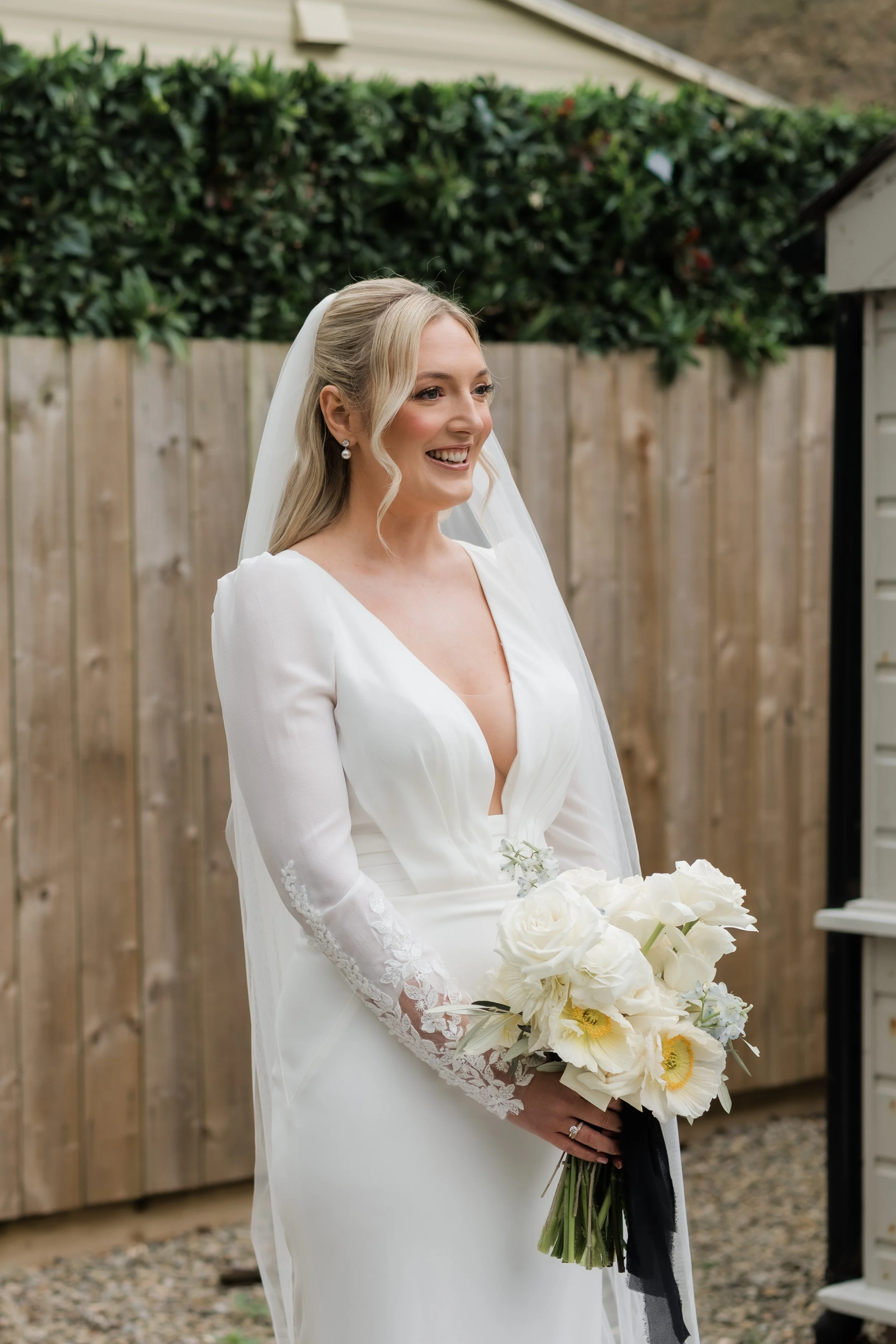 A bride in a white wedding dress holding a bouquet of white flowers, standing outdoors in front of a wooden fence with greenery.