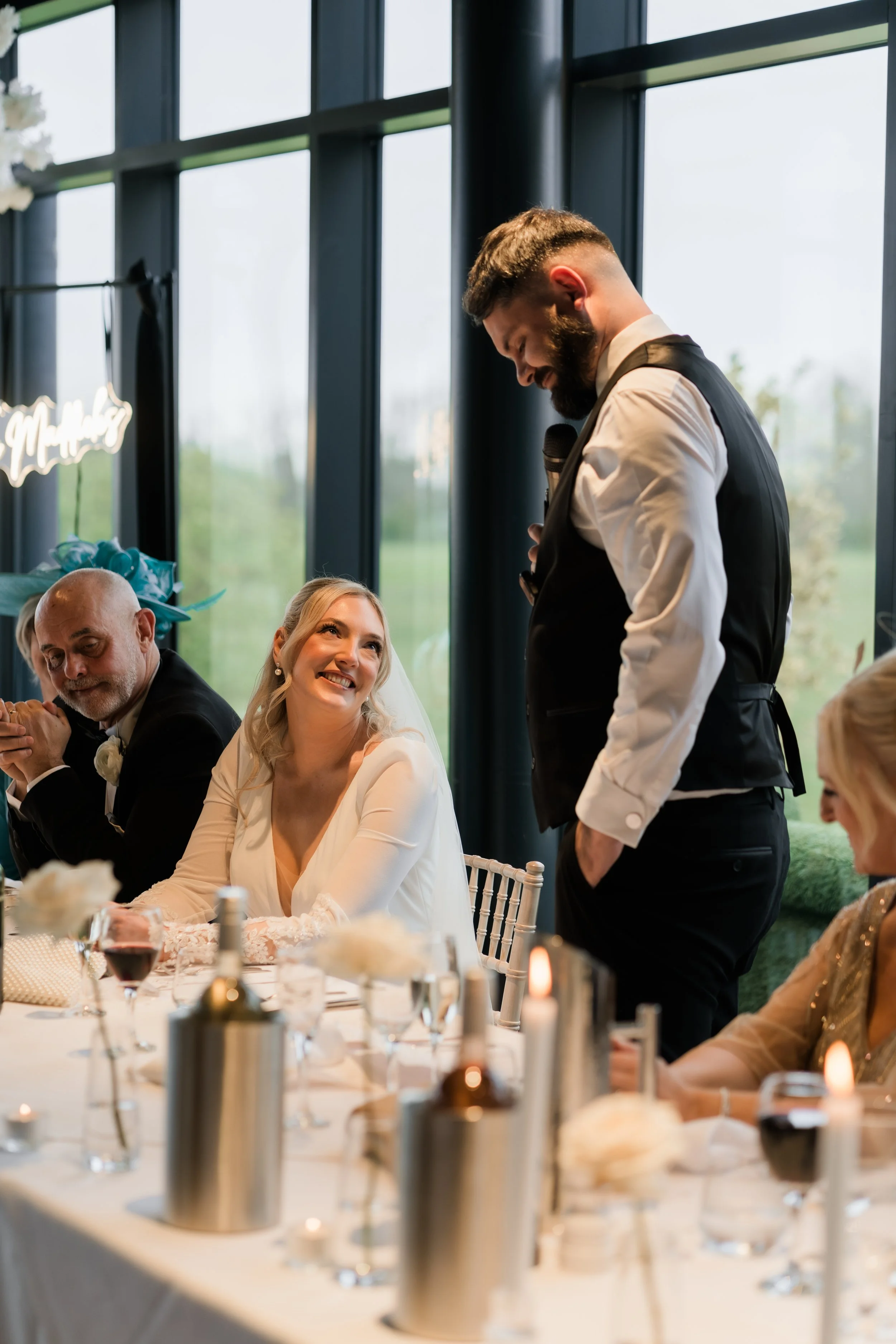 A man giving a speech at a wedding reception, standing next to the bride and guests seated at a decorated table with candles and wine, in a brightly lit room with large windows showing greenery outside.