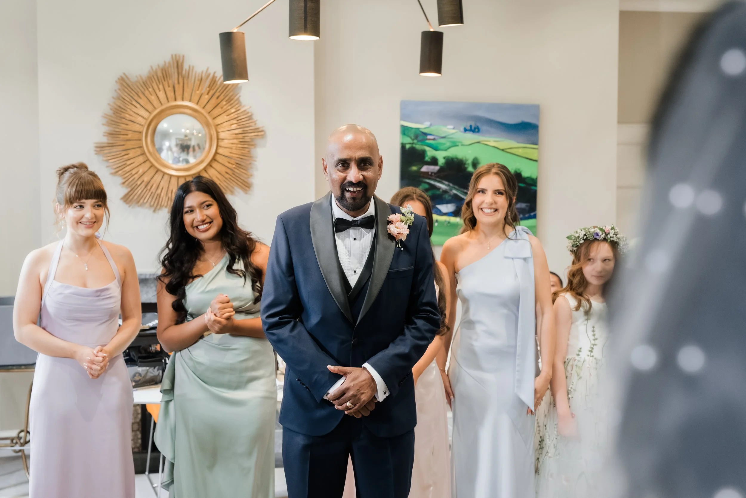 A groom in a dark blue tuxedo with a boutonniere, smiling at a wedding reception, surrounded by bridesmaids and flower girls in elegant dresses, with a decorative wall mirror and colorful painting in the background.