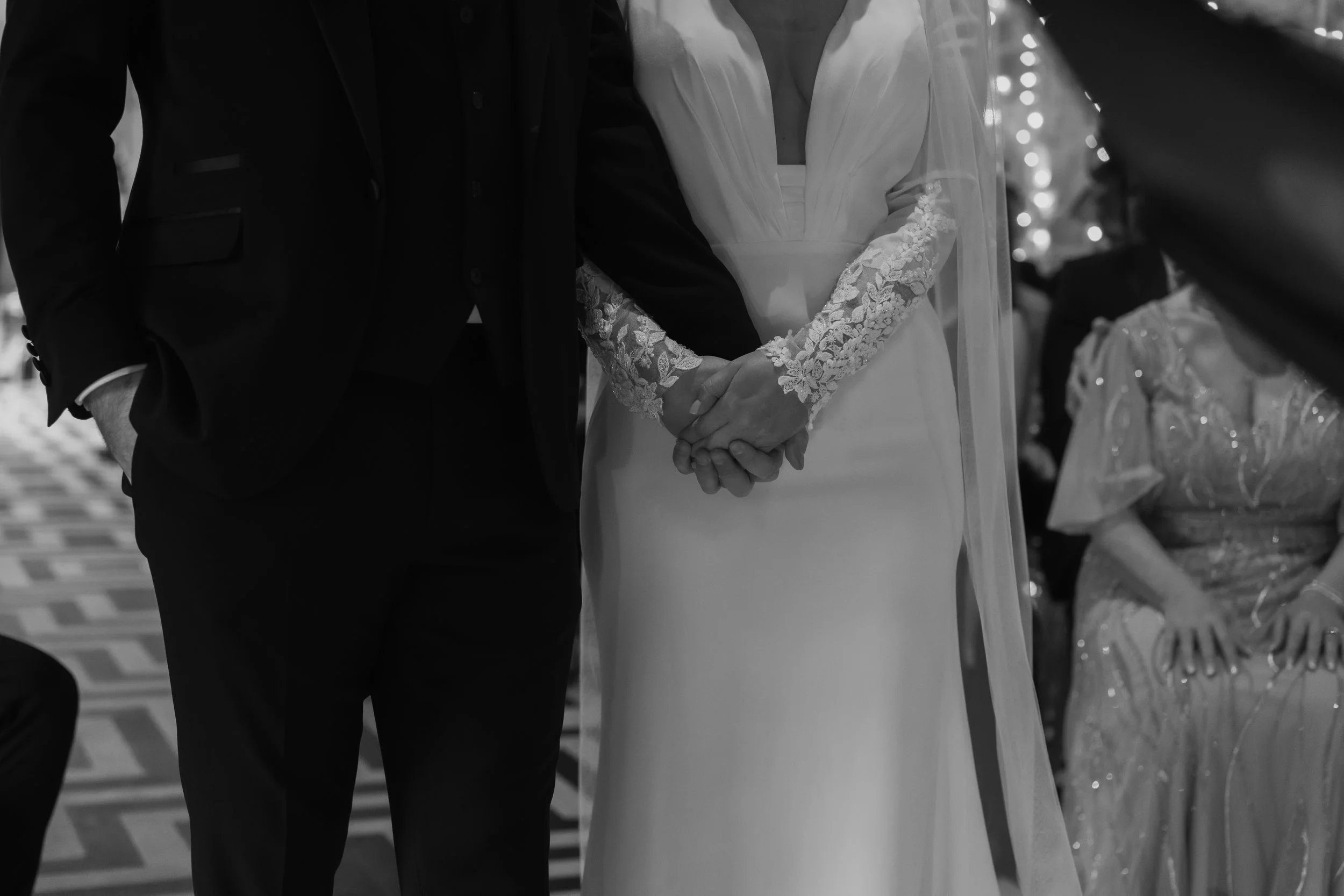 A black and white photo of a bride and groom holding hands during a wedding ceremony. The bride is wearing a gown with lace sleeves and the groom is in a suit.