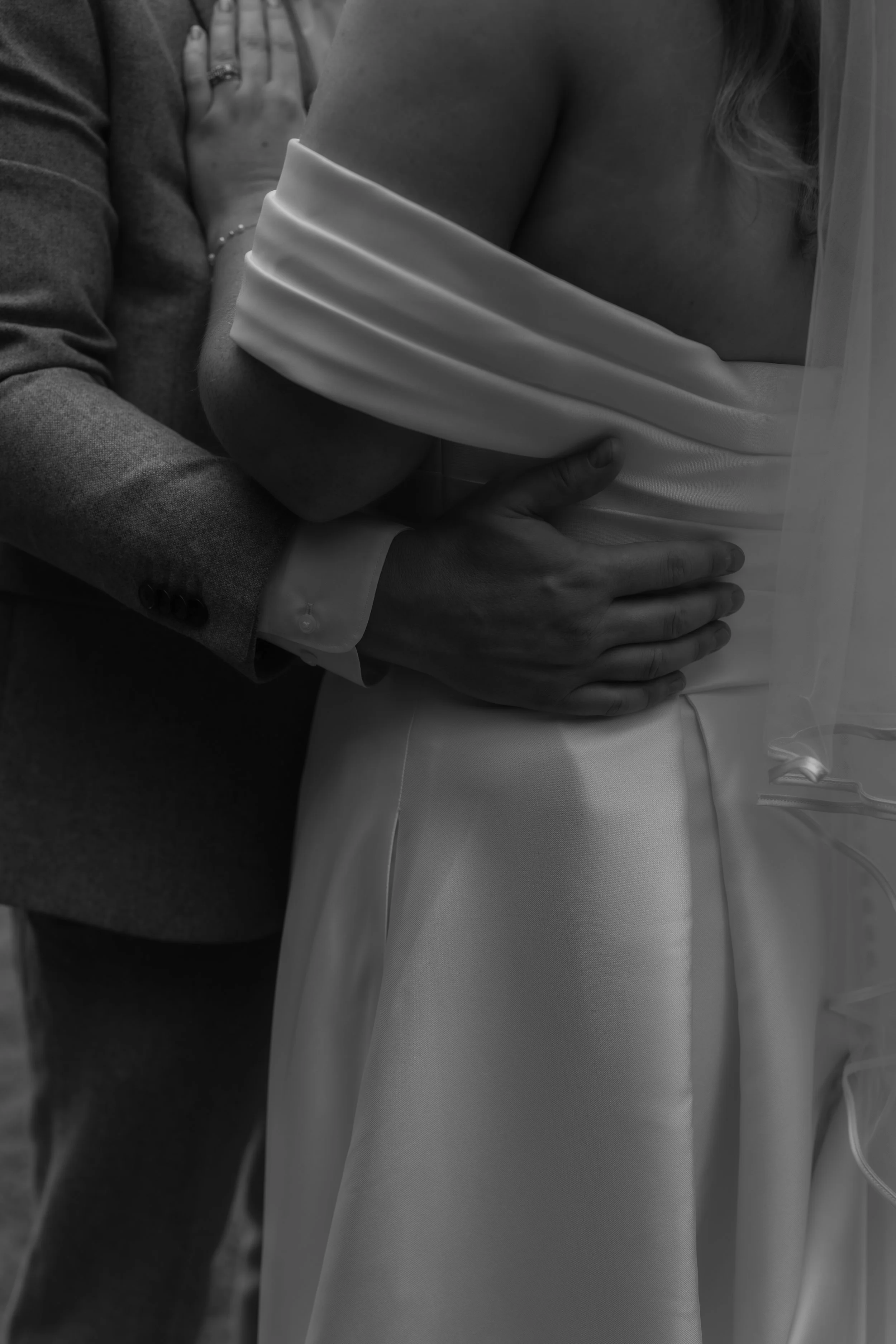 A close-up black-and-white photo of a couple's embrace, with a man’s arm around a woman’s waist, both wearing wedding attire.