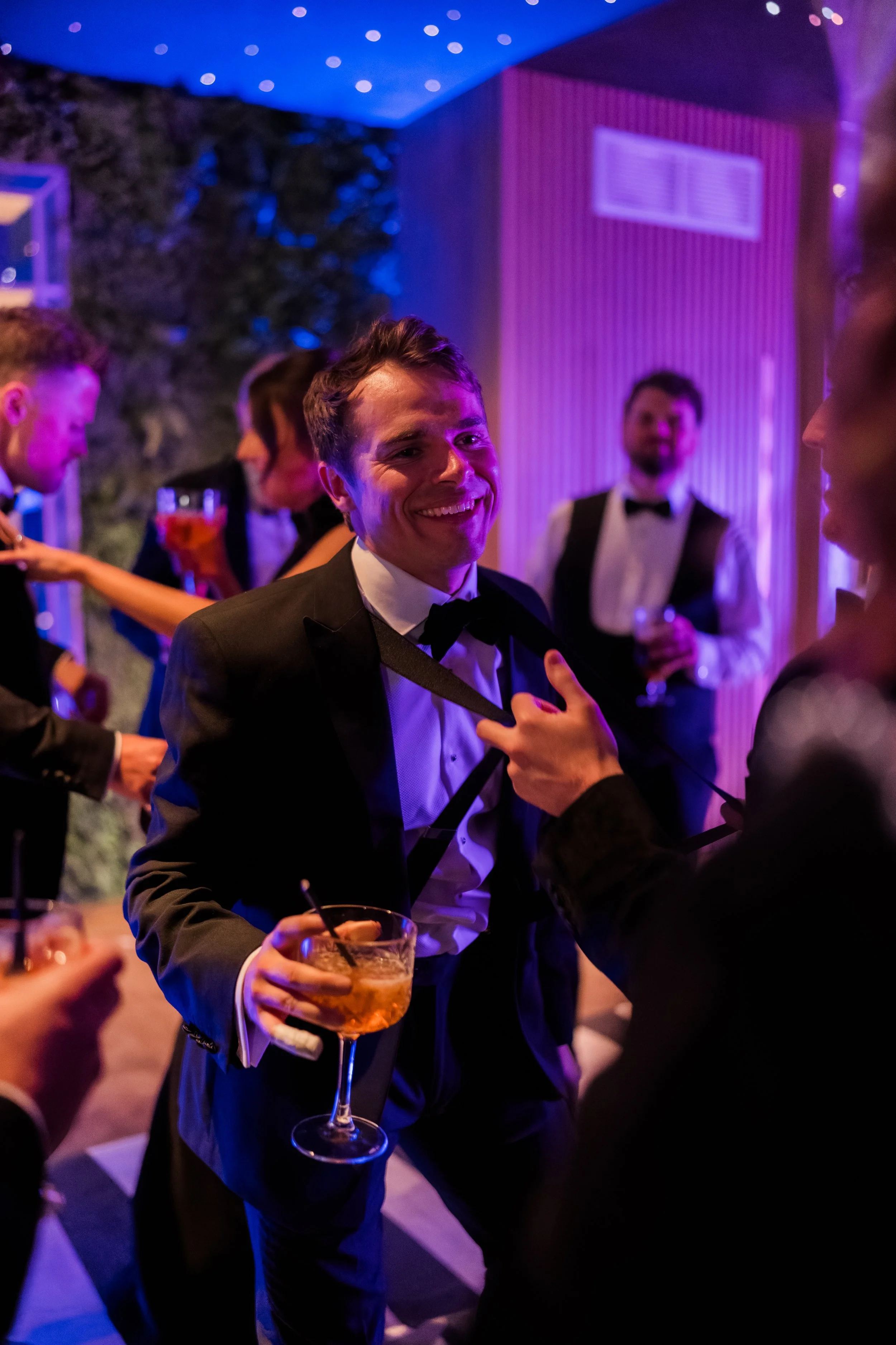 People in tuxedos dancing and socializing at a party with colorful lighting and a stone wall in the background.