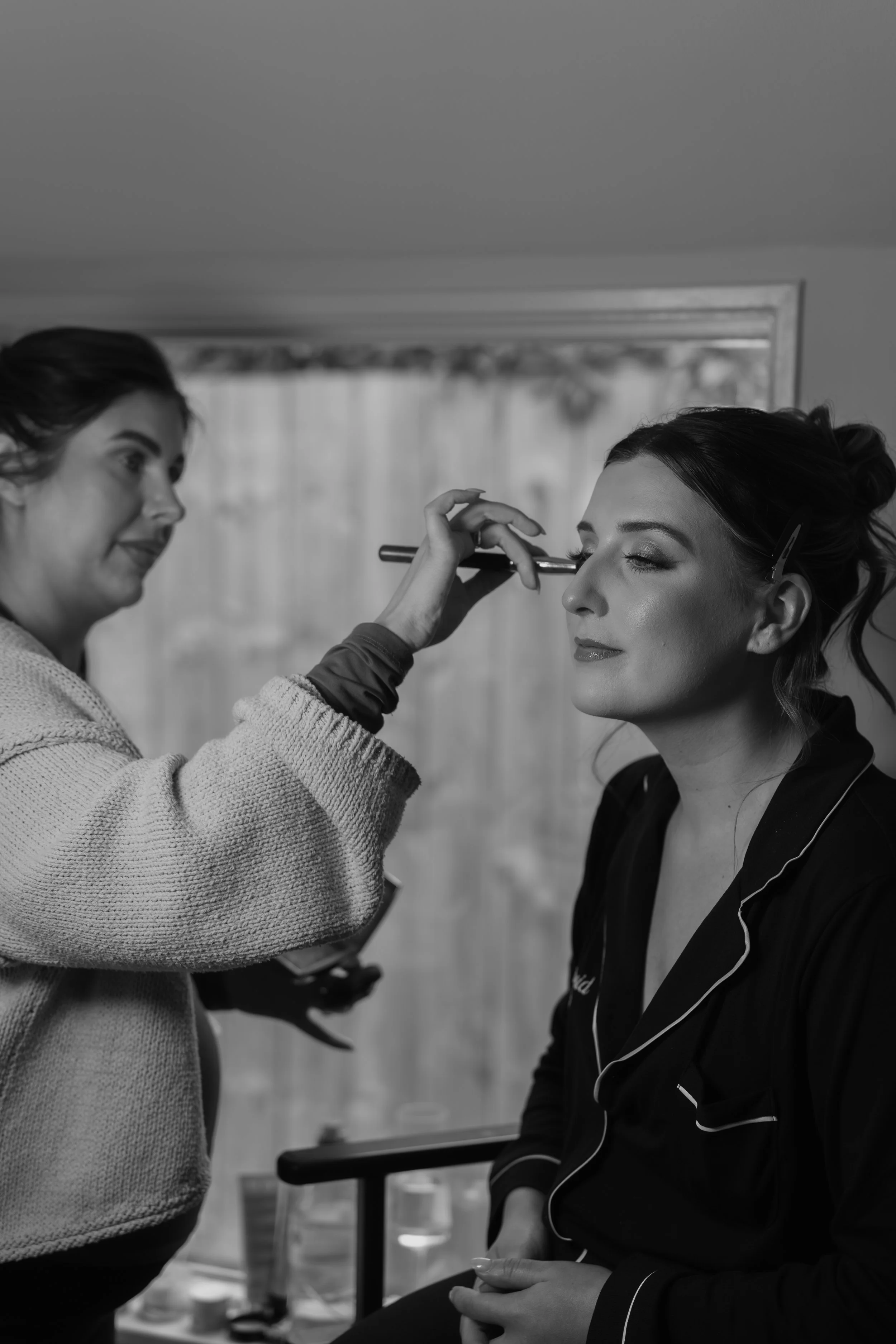 A woman in pajamas is getting her makeup done by a makeup artist with a brush on her eyelid. The woman is seated with her eyes closed and has her hair styled.