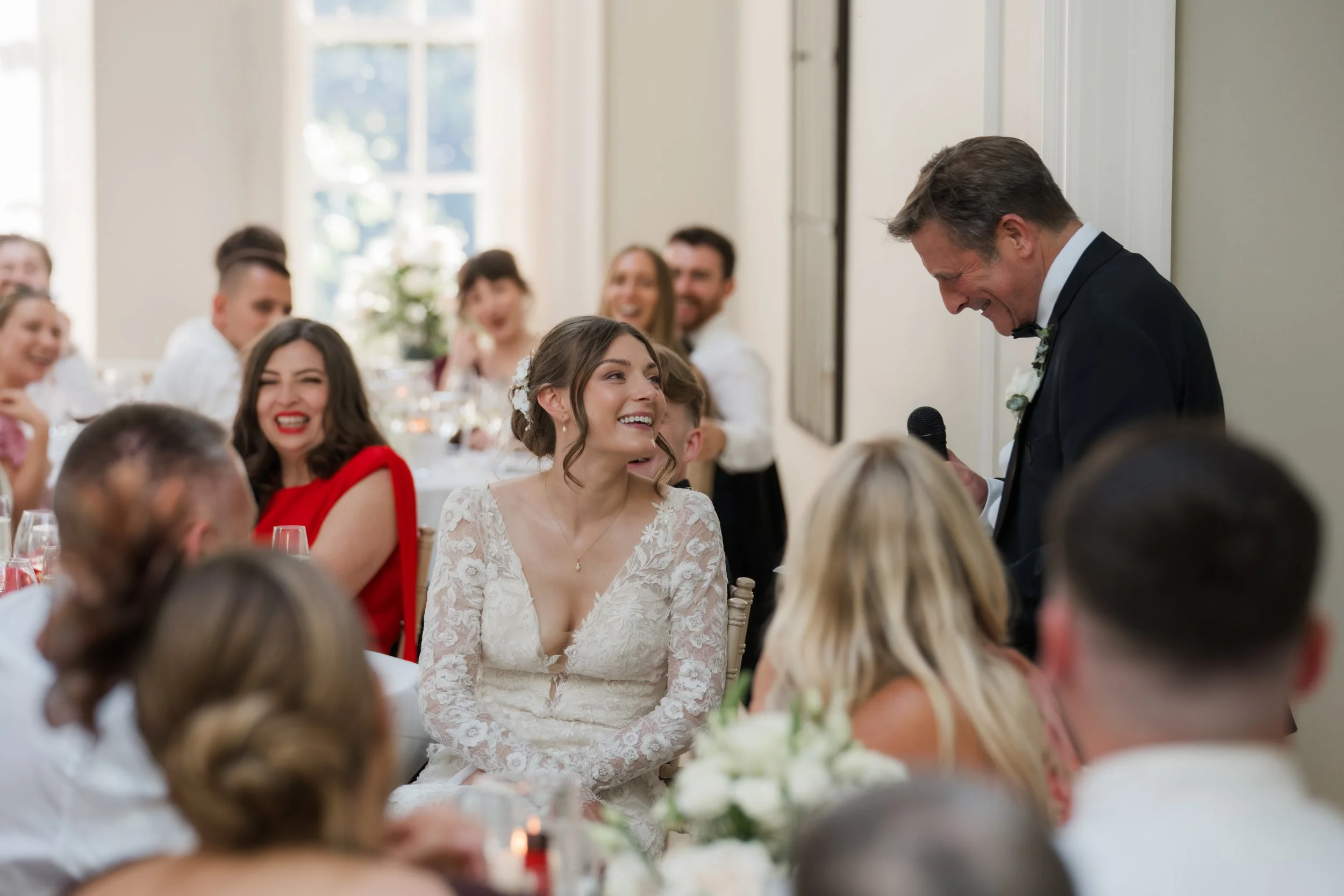 Bride in a lace wedding gown sitting at a wedding reception table, smiling and looking at an older man in a suit who is holding a microphone. Other guests are seated around the table, some smiling and applauding. The room is bright with natural light