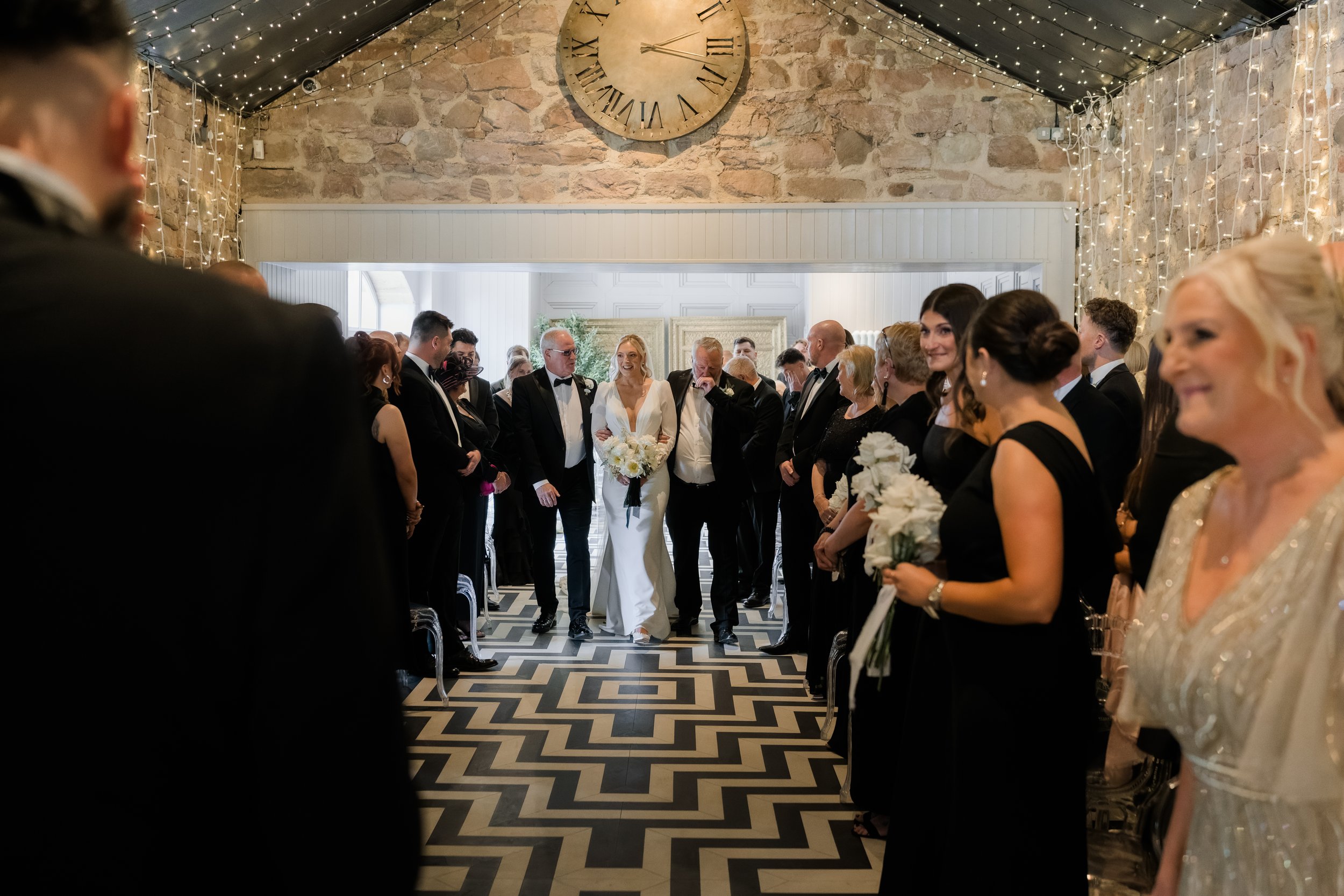 A bride walking down the aisle with her parents during a wedding ceremony, surrounded by guests in formal attire inside a decorated venue with string lights and a large clock on the wall.