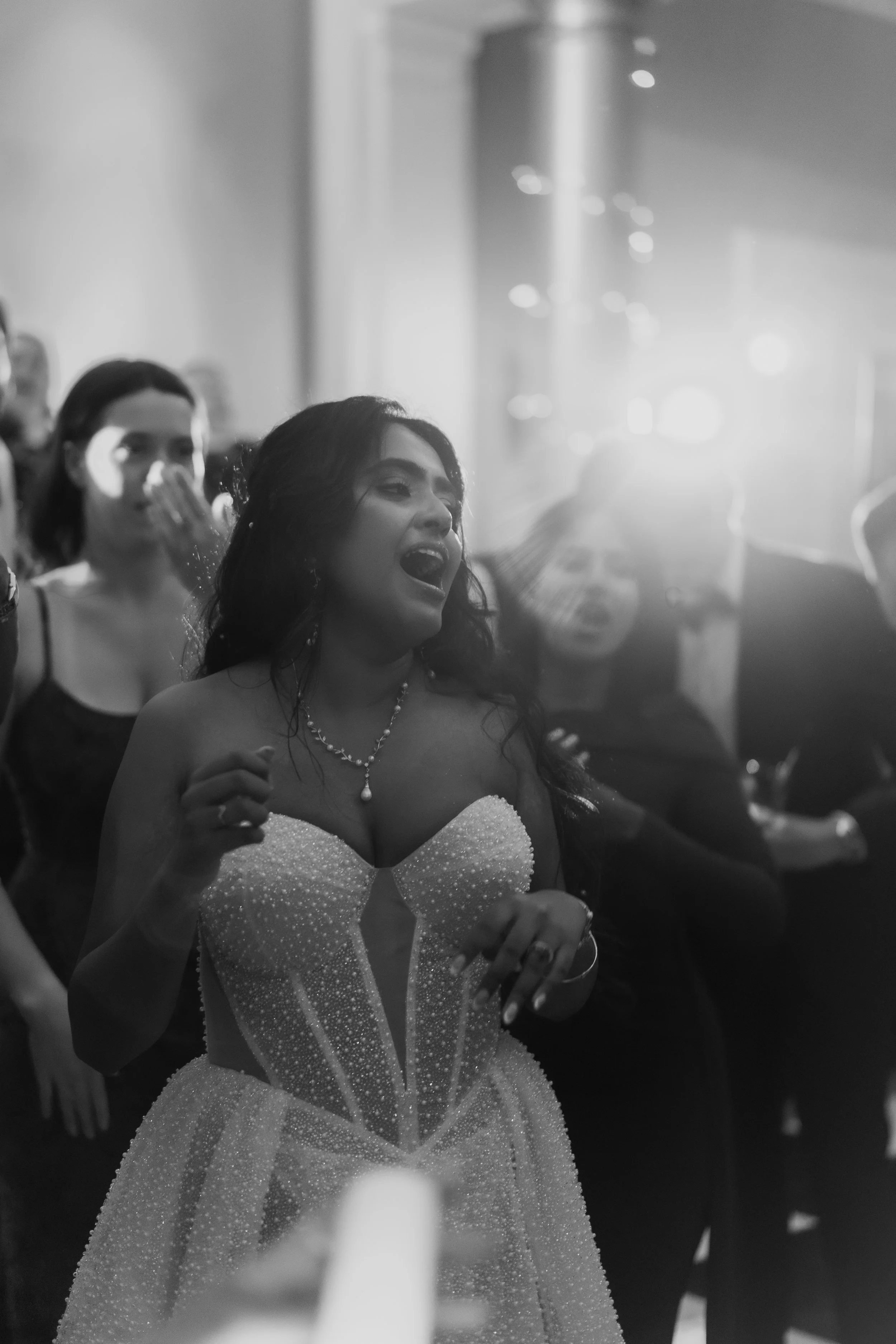 A woman in an elegant strapless dress singing or speaking passionately at a formal event, surrounded by other guests who are listening or reacting.