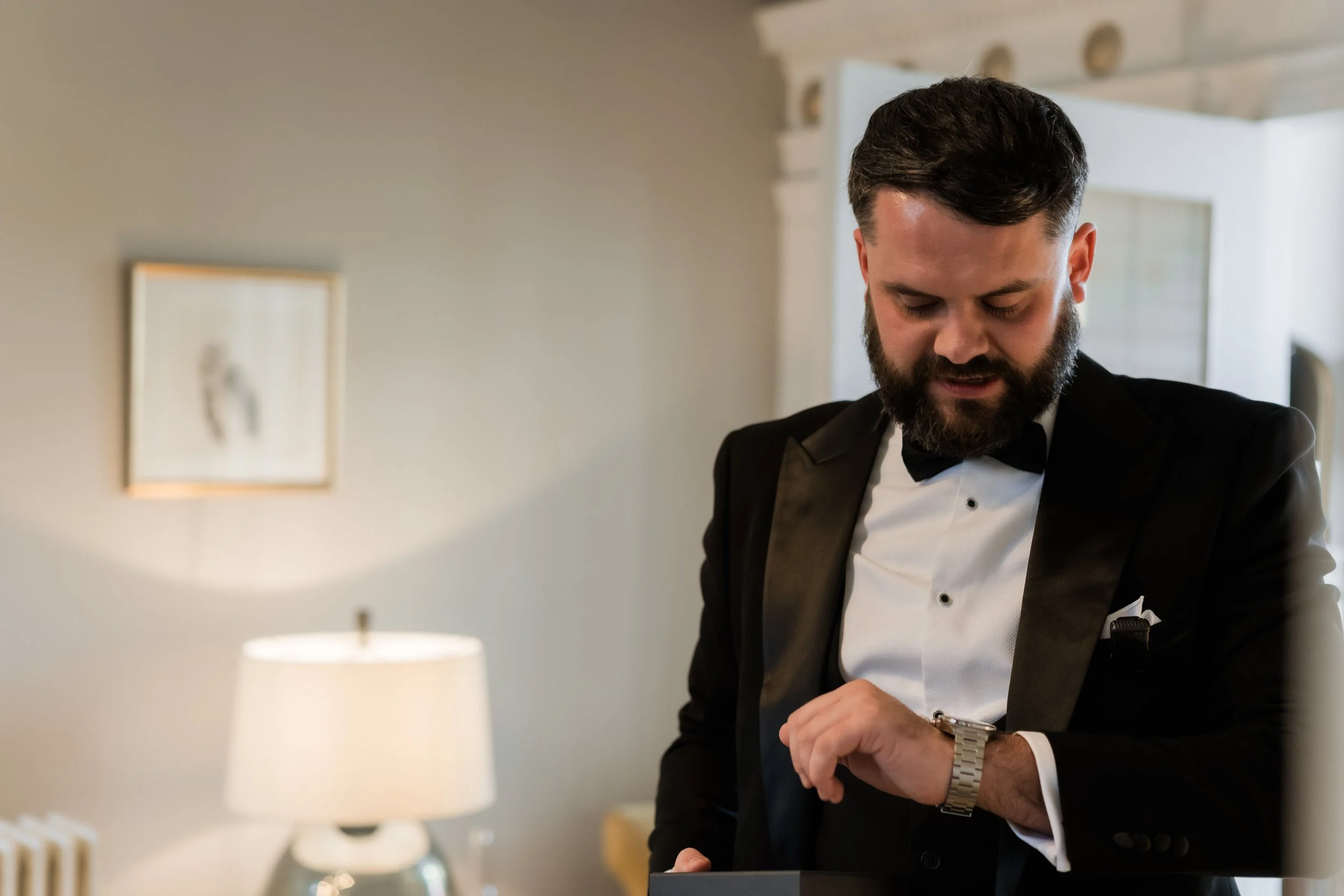 A man in a tuxedo looking at his watch in a softly lit room.