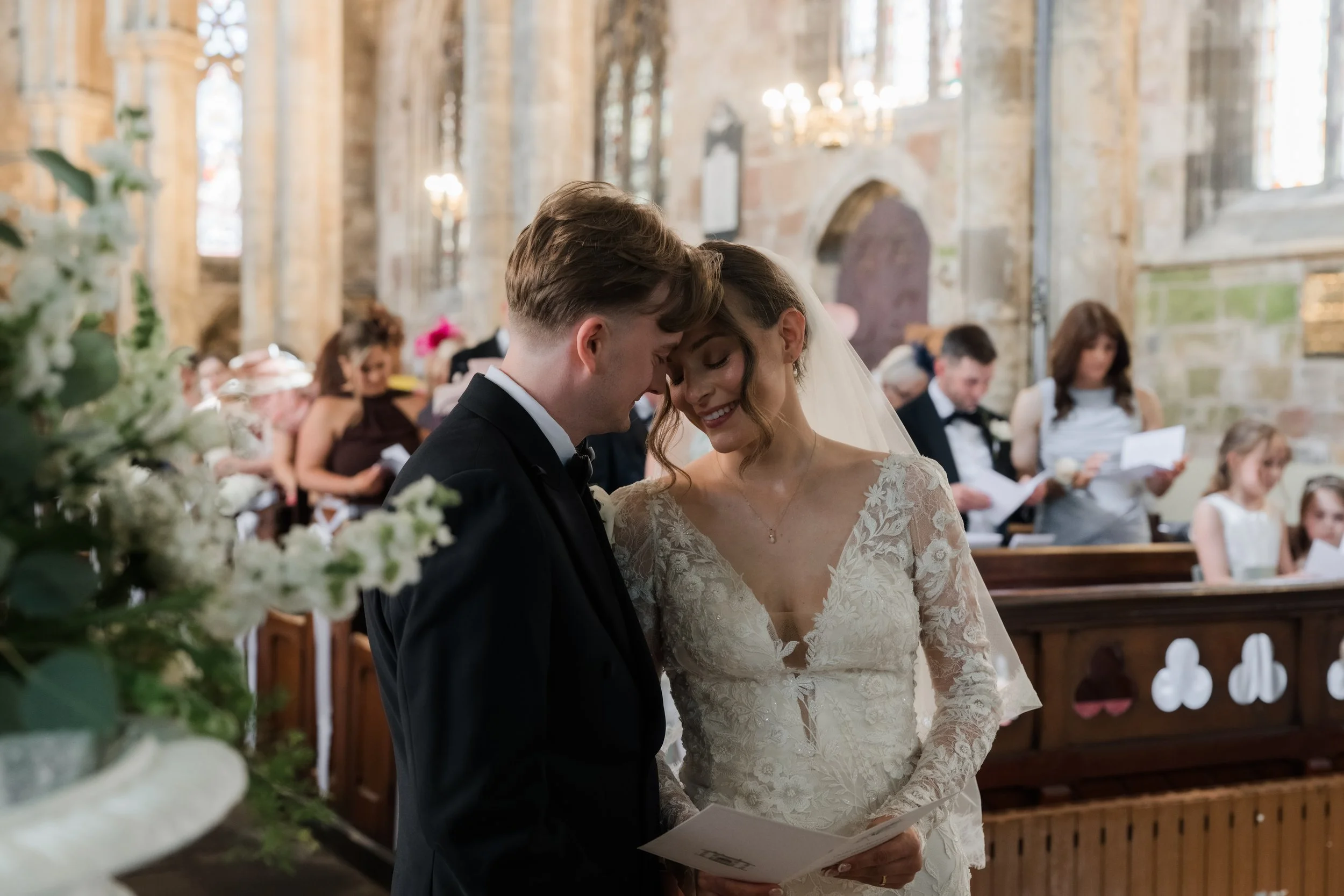 A bride and groom standing close together during their wedding ceremony inside a church, with guests seated behind them.