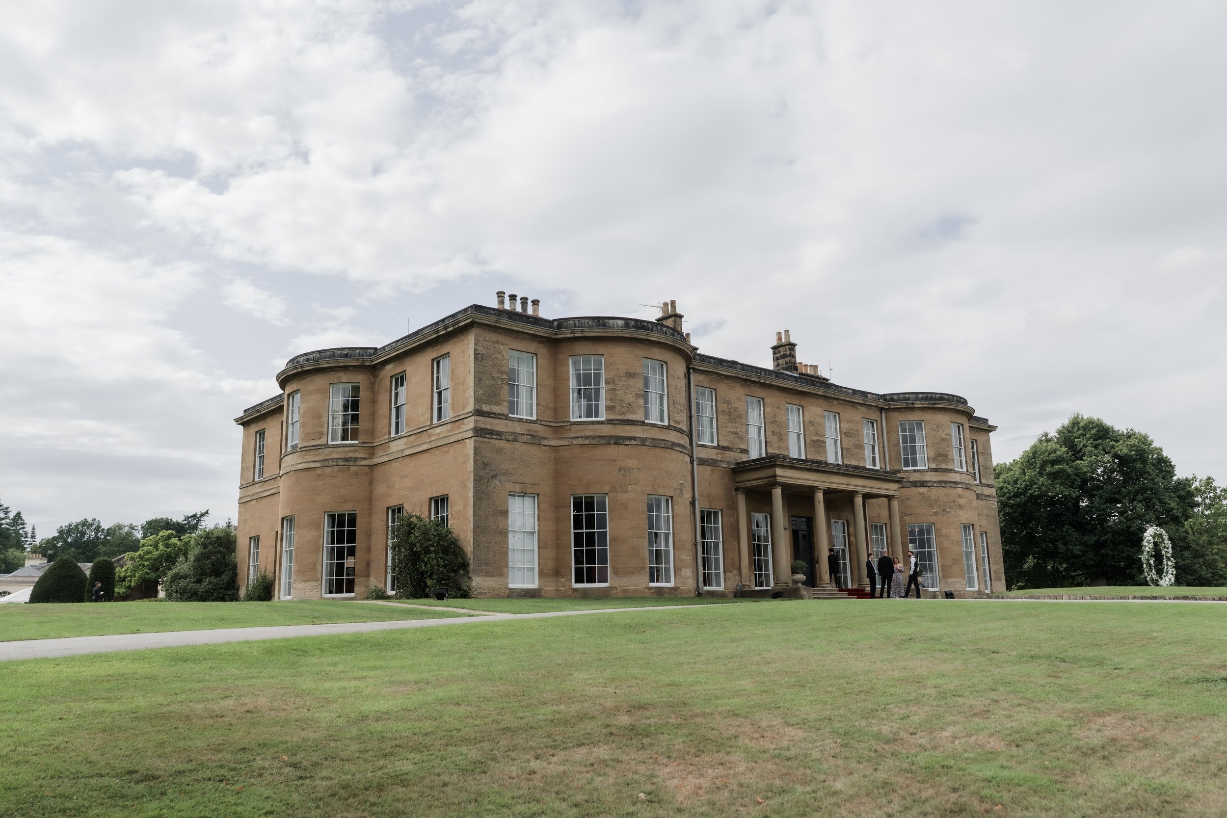 A large, historic mansion with beige stone walls, multiple tall windows, and a front porch with columns. Several people are standing near the entrance on a grassy lawn under a cloudy sky.