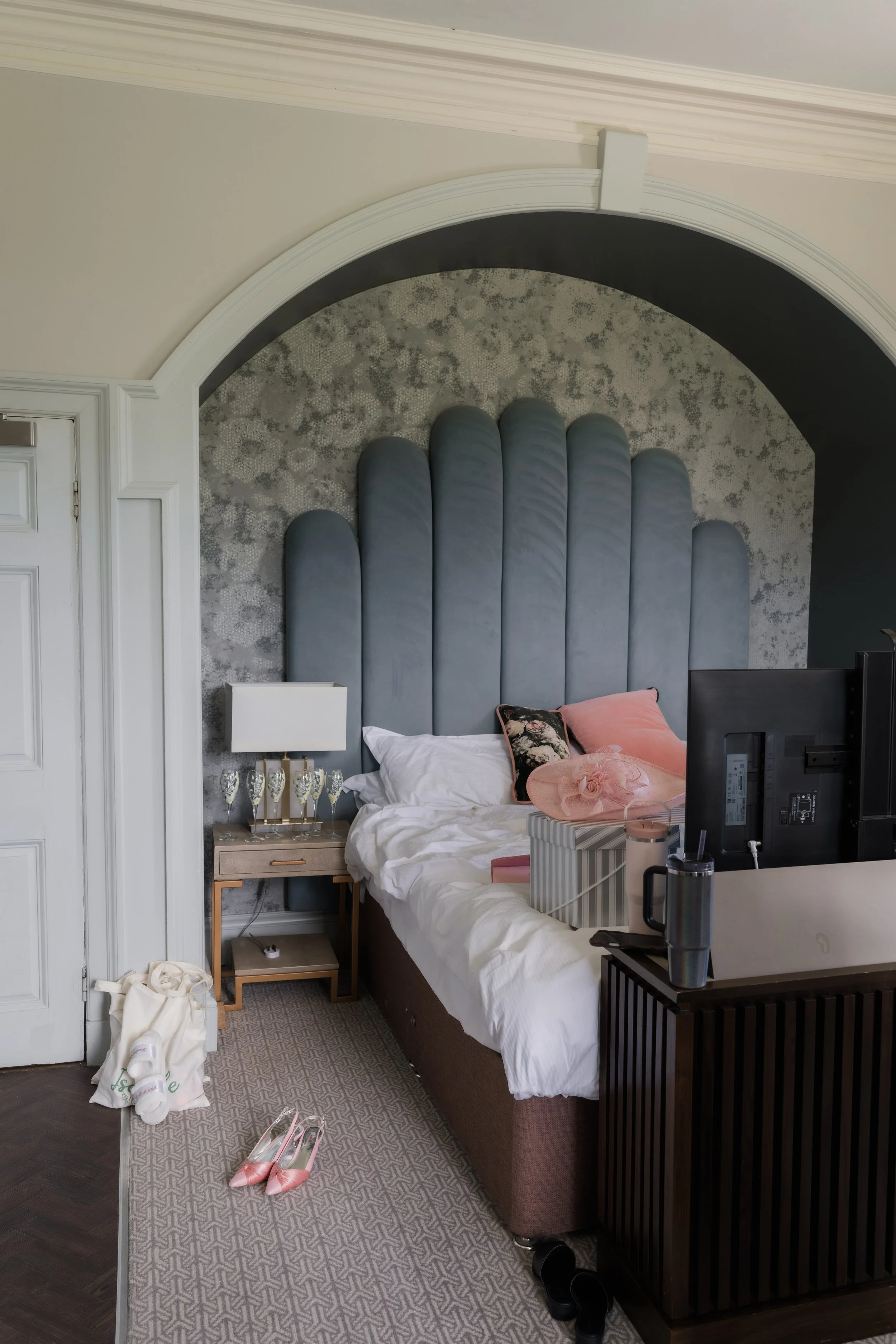 A bedroom with a bed, pillows, and a bedside table with glasses of champagne. There are pink high heels on the rug and a white tote bag on the floor. The headboard is padded and tall, with an arch above it, and there's a decorative wall behind with t