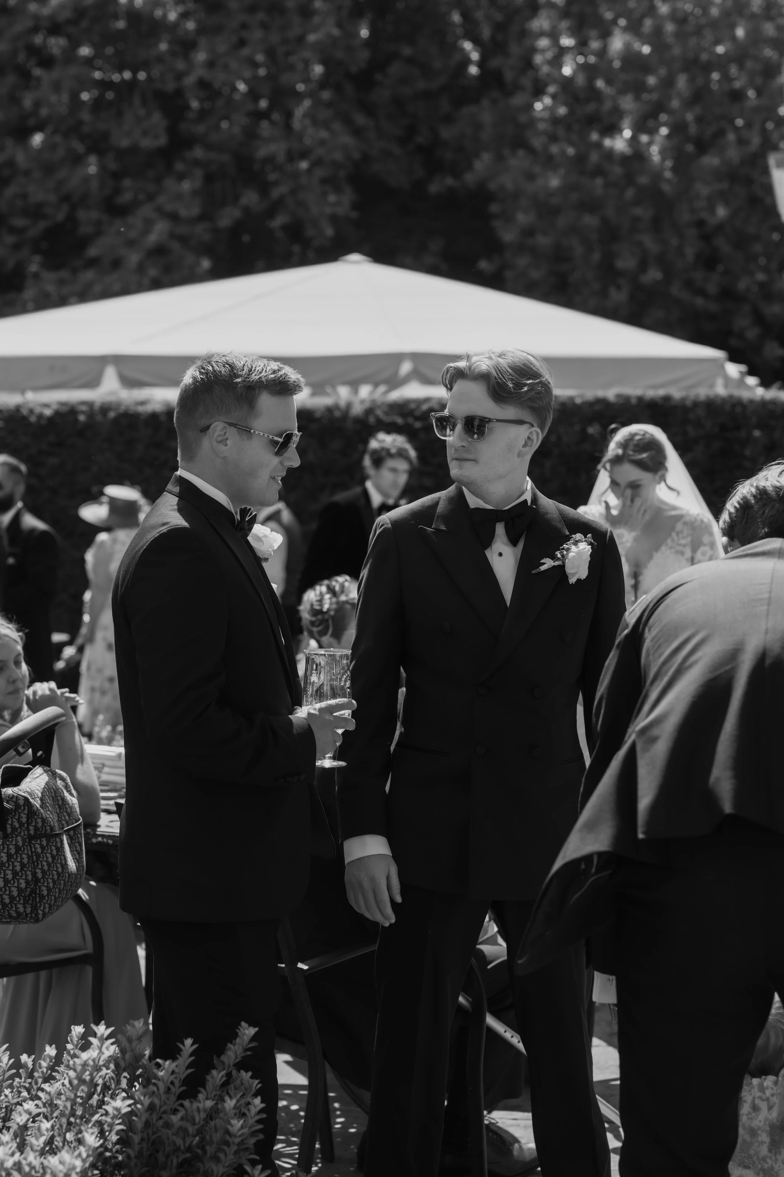 Black and white photo of two men in tuxedos and sunglasses at a wedding reception outdoors, drinking champagne, with guests and a tent in the background.