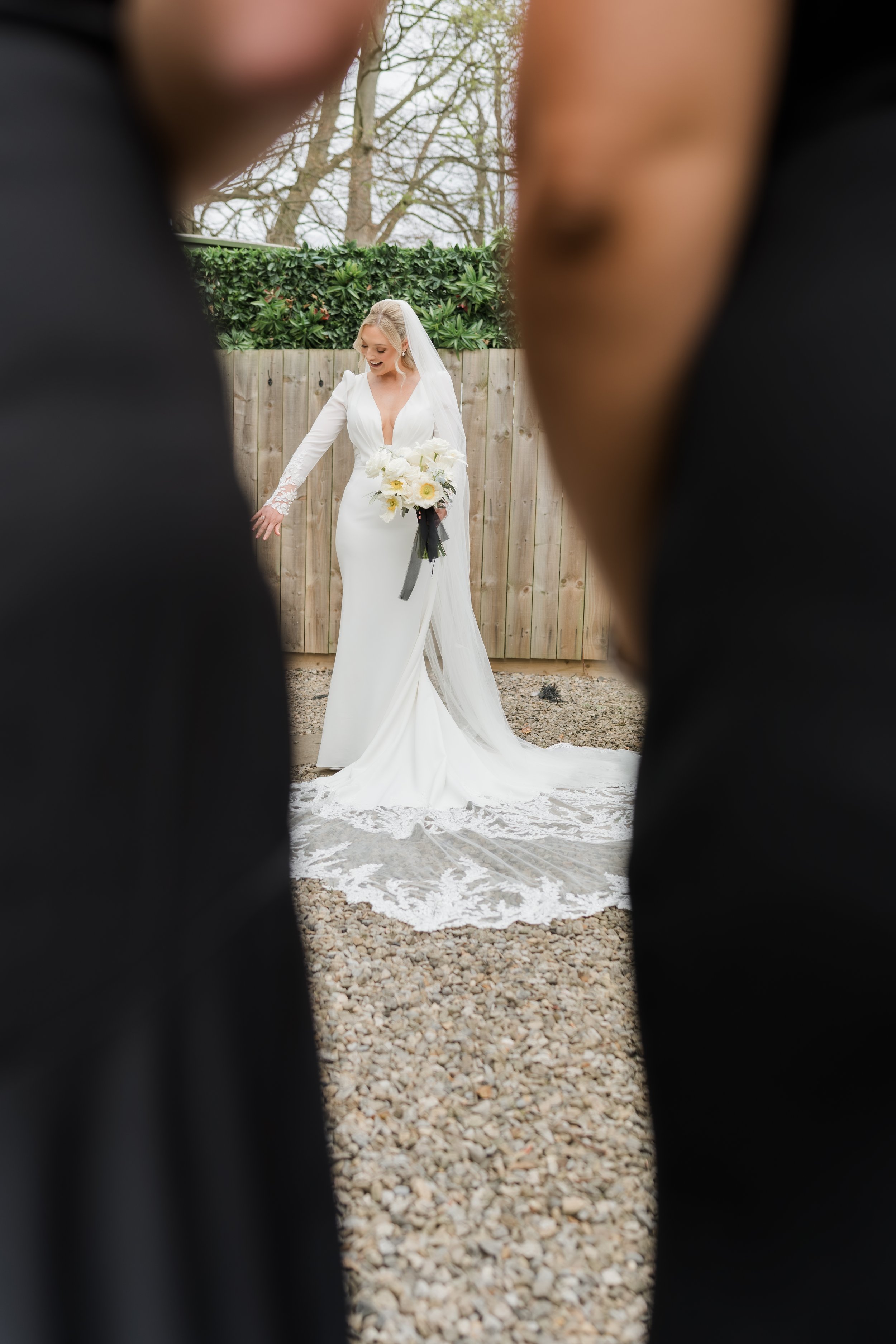 Bride in a white wedding dress holding a bouquet, seen through an opening between two people dressed in black.