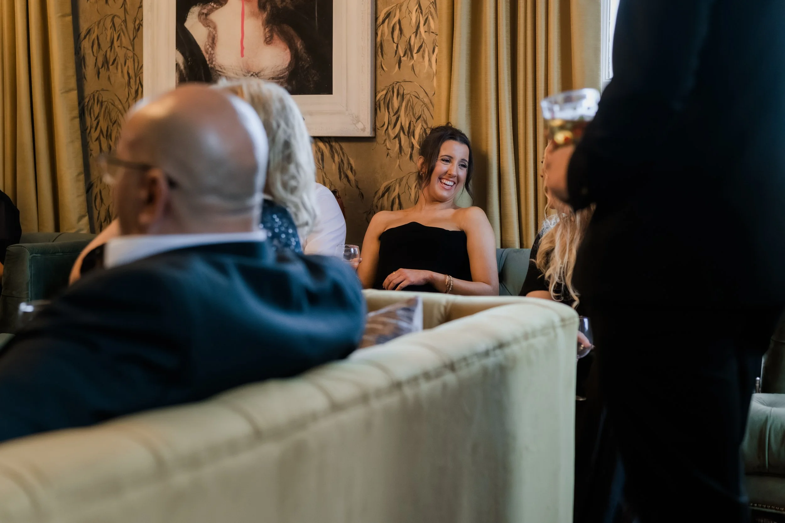 A group of people at a social gathering, with a woman in a black dress smiling and laughing, seated on a green sofa, while others stand and sit around her in a warmly decorated room.
