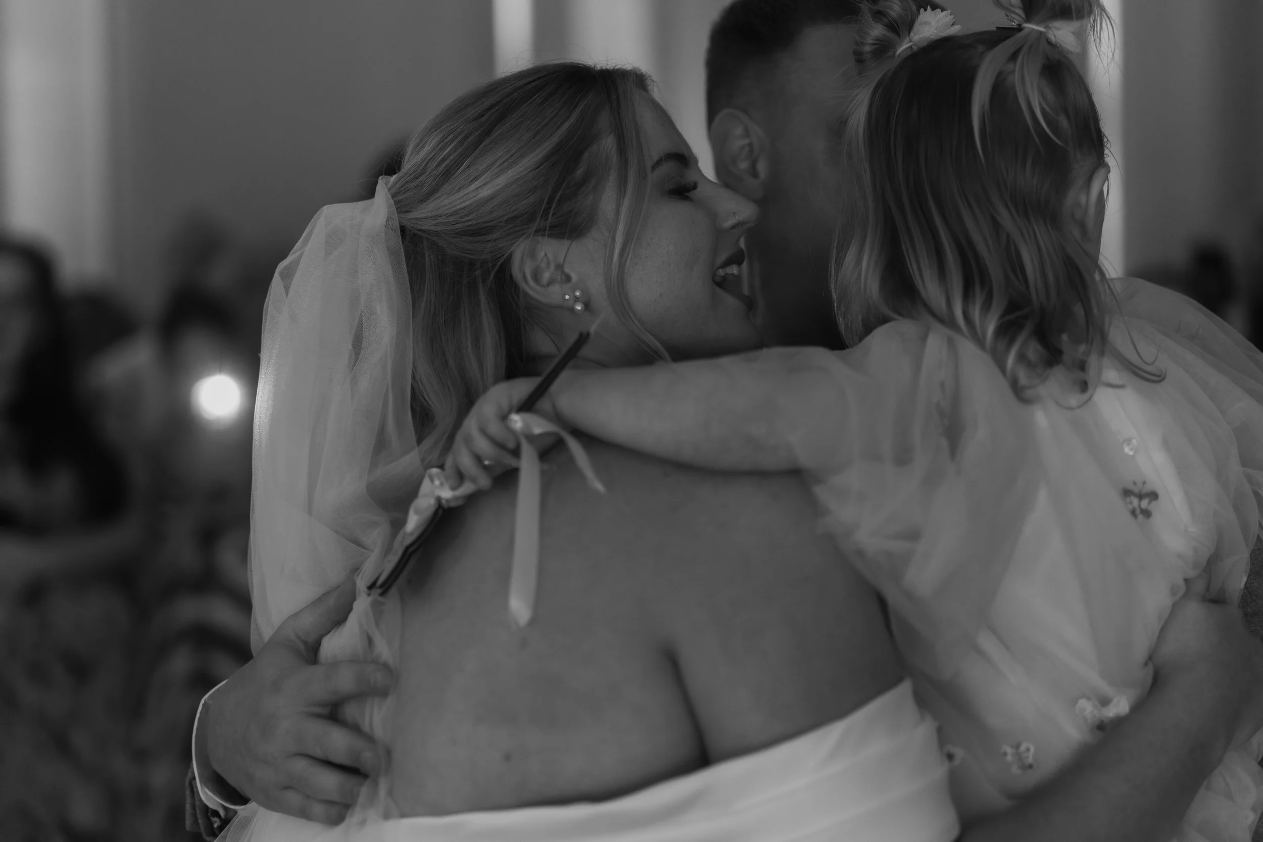 A woman in a wedding dress hugging two children, one on each side, in a tender embrace at a wedding ceremony.