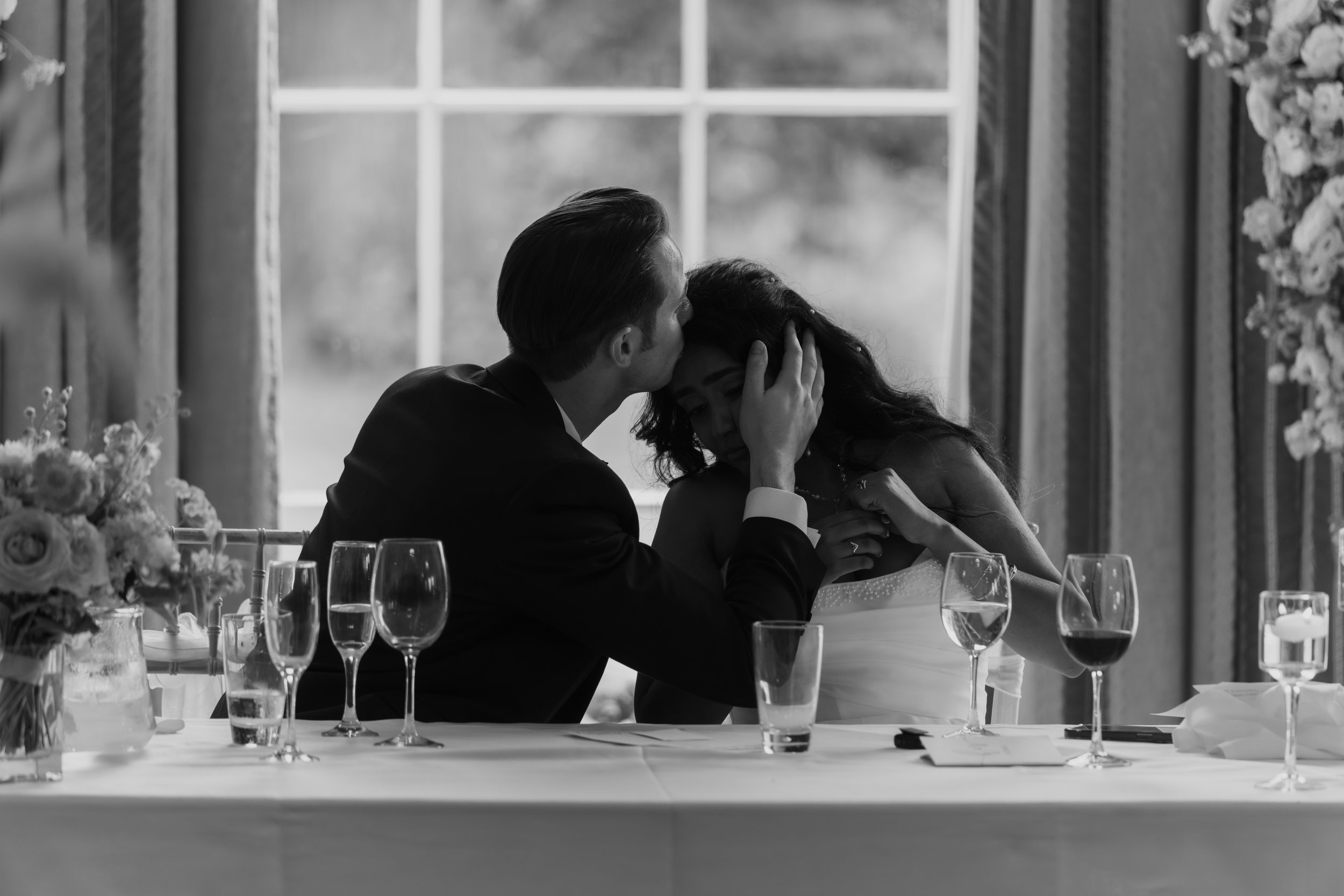 A black and white photo of a man and woman at a formal event, the man is comforting the woman who appears distressed, sitting at a table with wine glasses and floral arrangements, near a large window.
