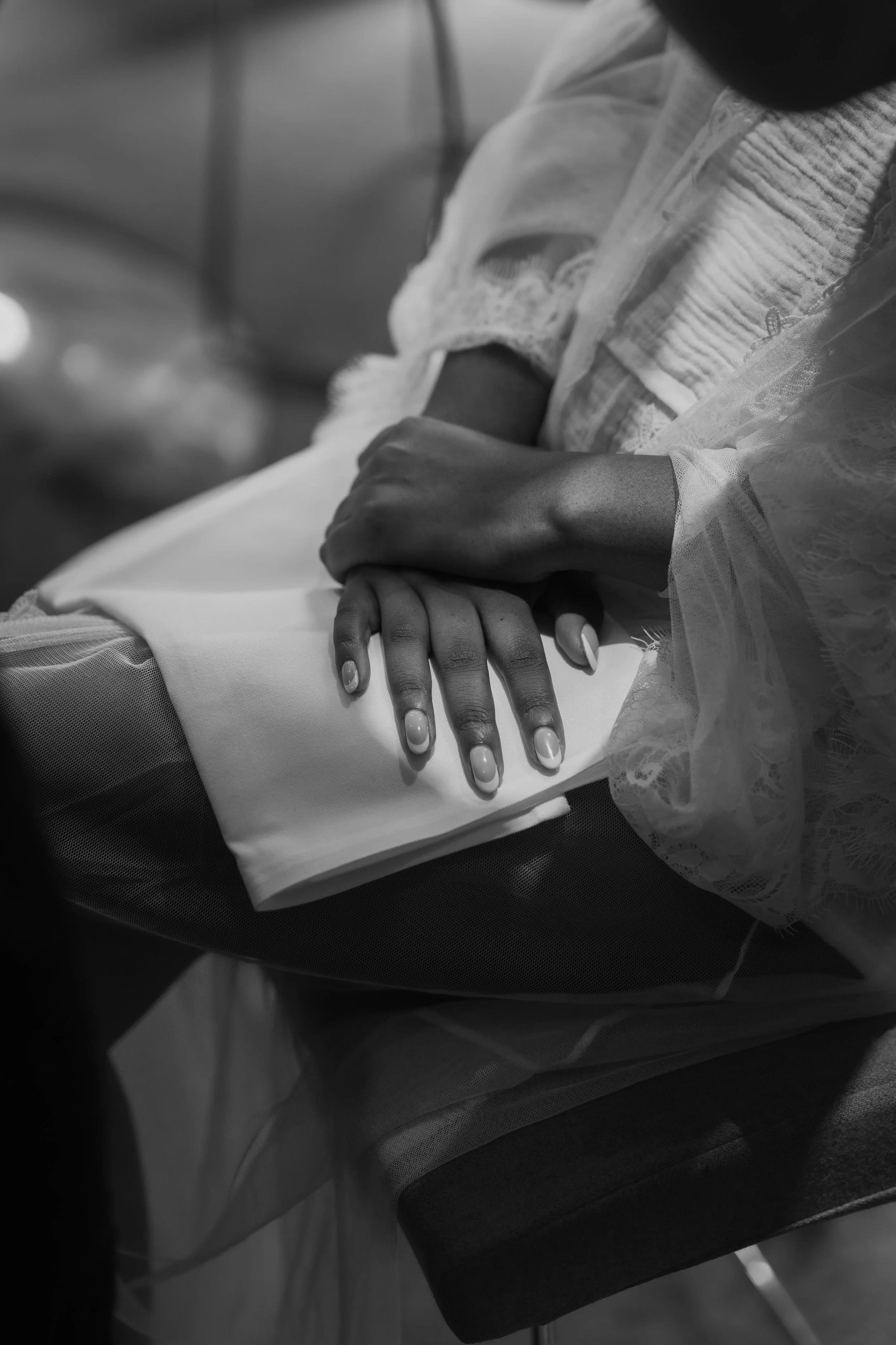 Close-up of a person's hands resting on their lap, with one hand overlapping the other, seated in a chair. The person is dressed in lace and light-colored clothing, with the photo taken in black and white.
