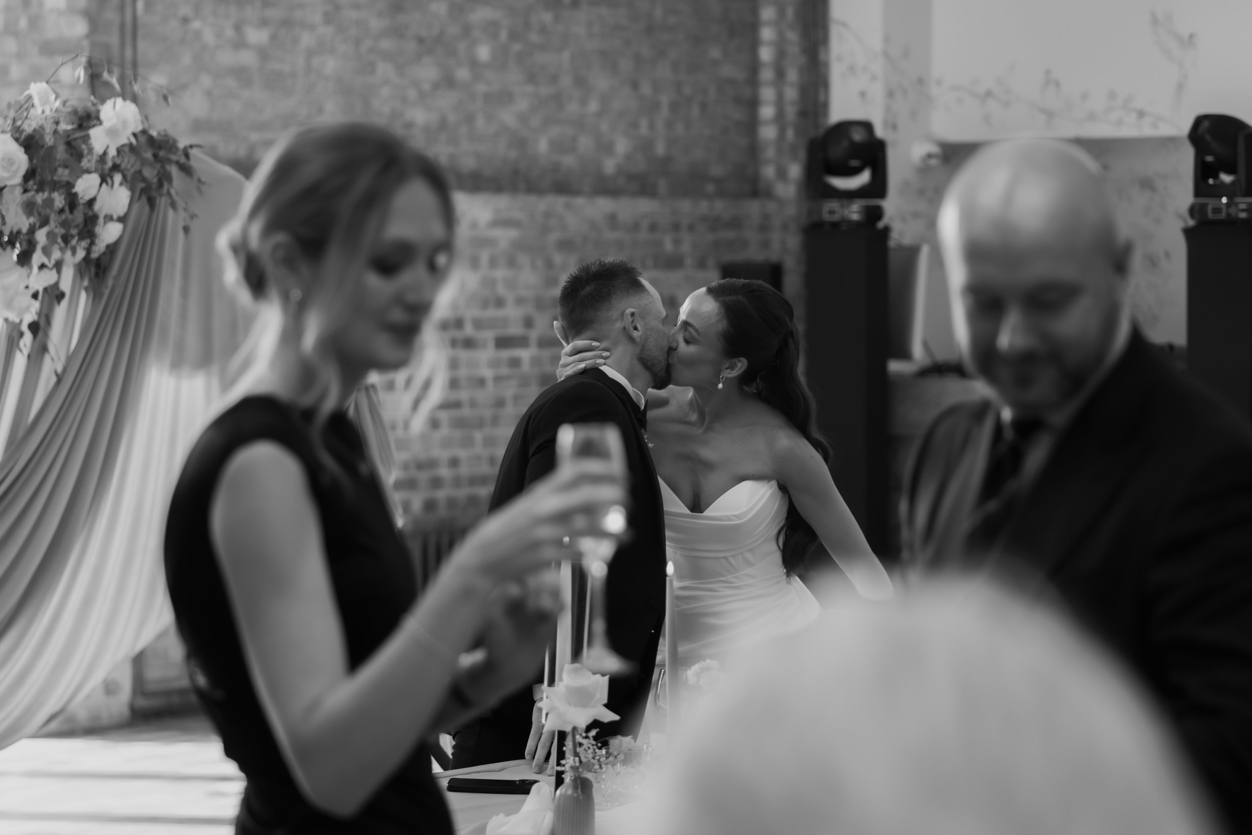 Black and white photo of a wedding reception with a bride and groom kissing, surrounded by guests seated at the dining table, with a floral centerpiece.
