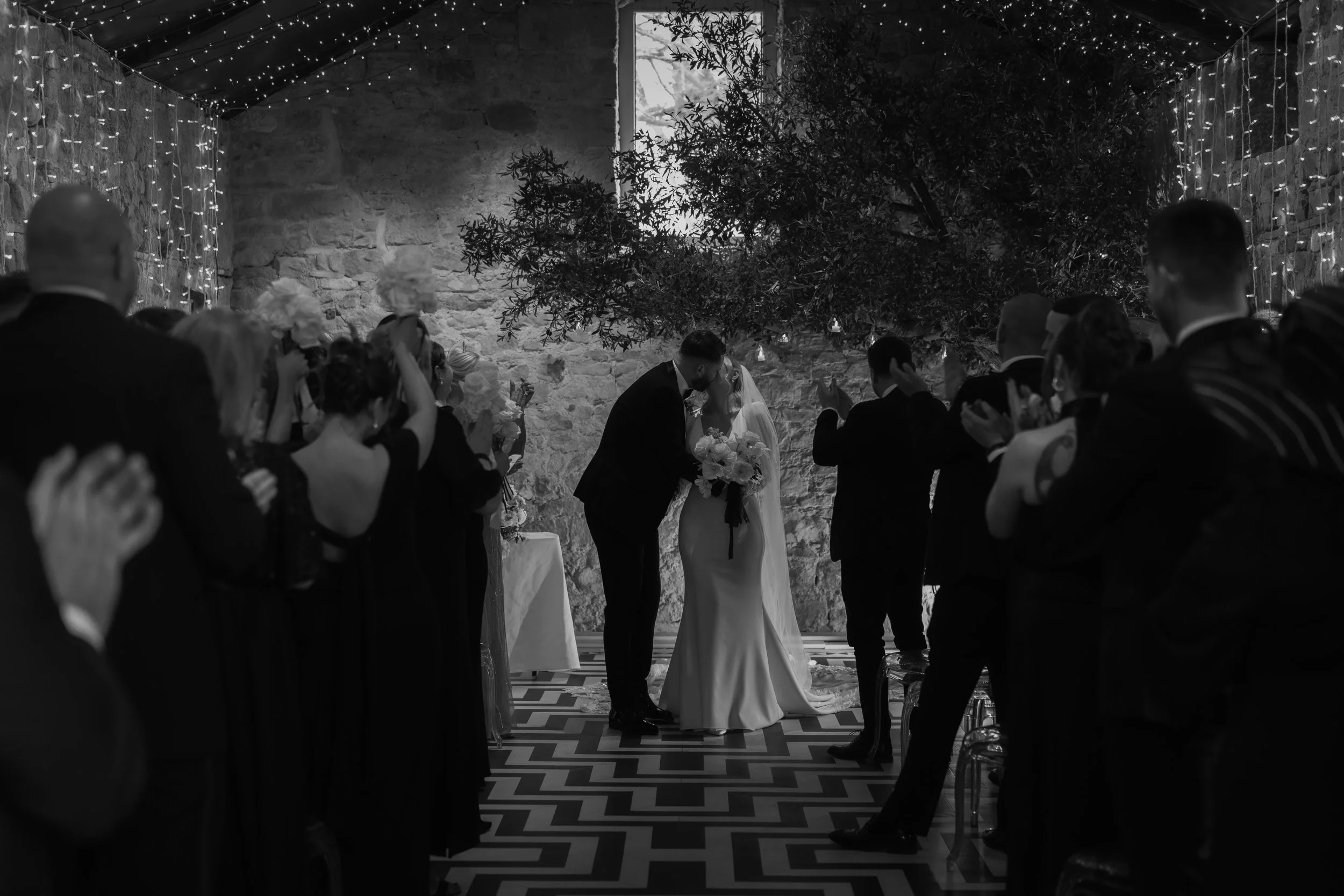 Black and white photo of a wedding ceremony with the bride and groom kissing, surrounded by guests.