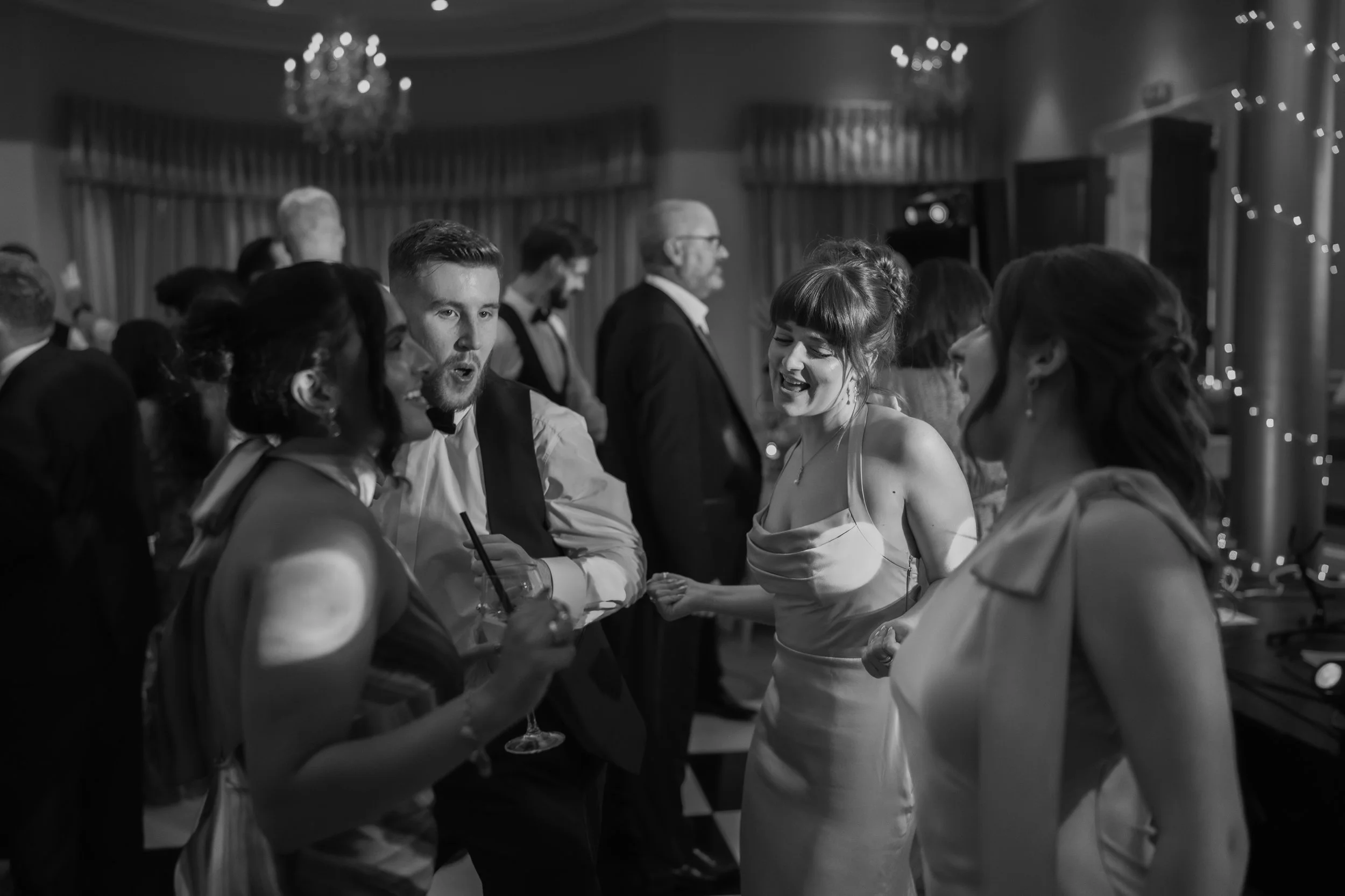 People dancing and talking at a formal social event, dressed in suits and elegant dresses, with a chandelier and string lights in the background.