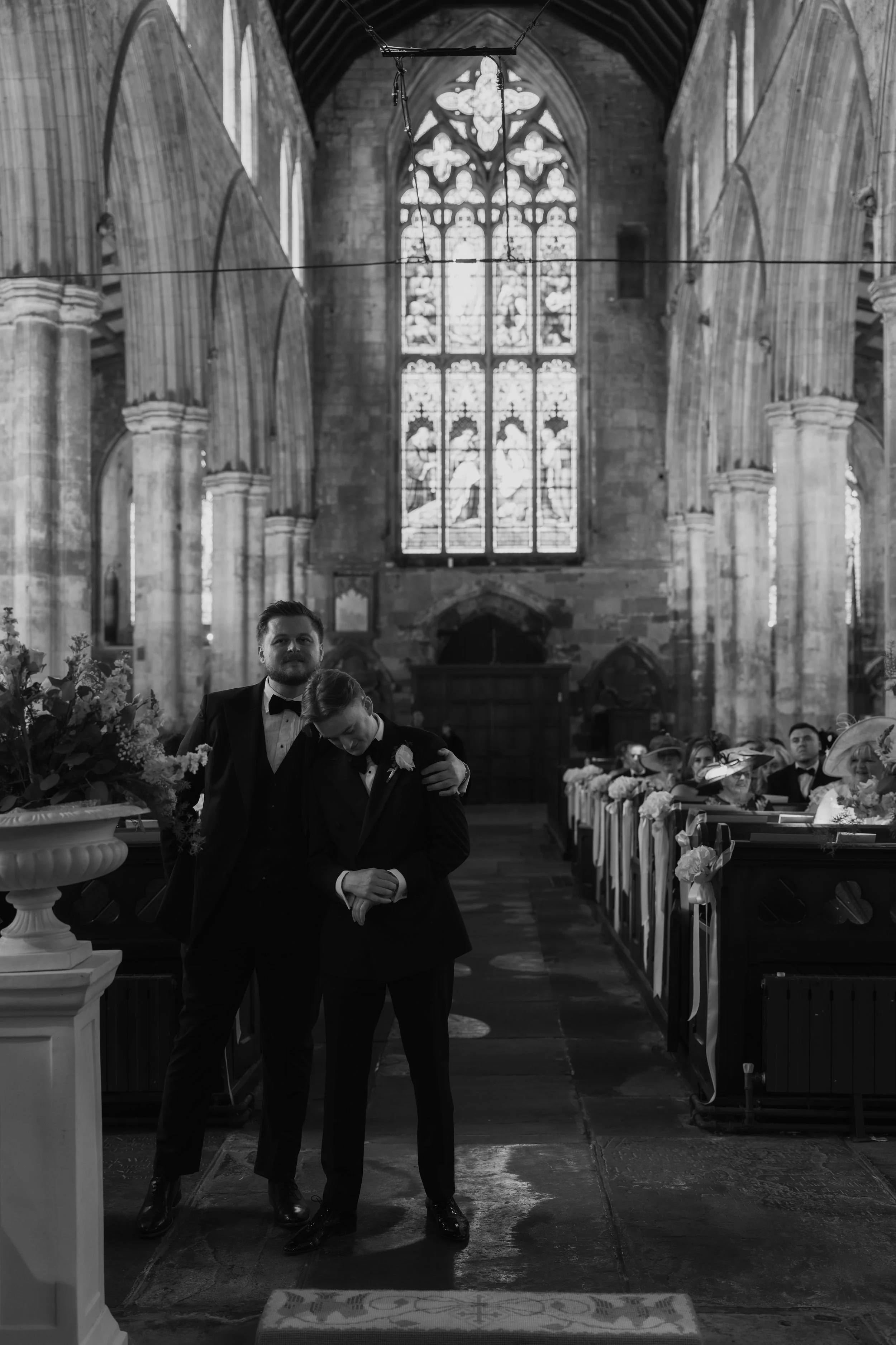 A black and white photo of a wedding ceremony inside a church with stained glass windows. In the foreground, a groom and bride are embracing. The groom is wearing a tuxedo with a bow tie. The bride is dressed in a wedding gown. Guests are seated on b