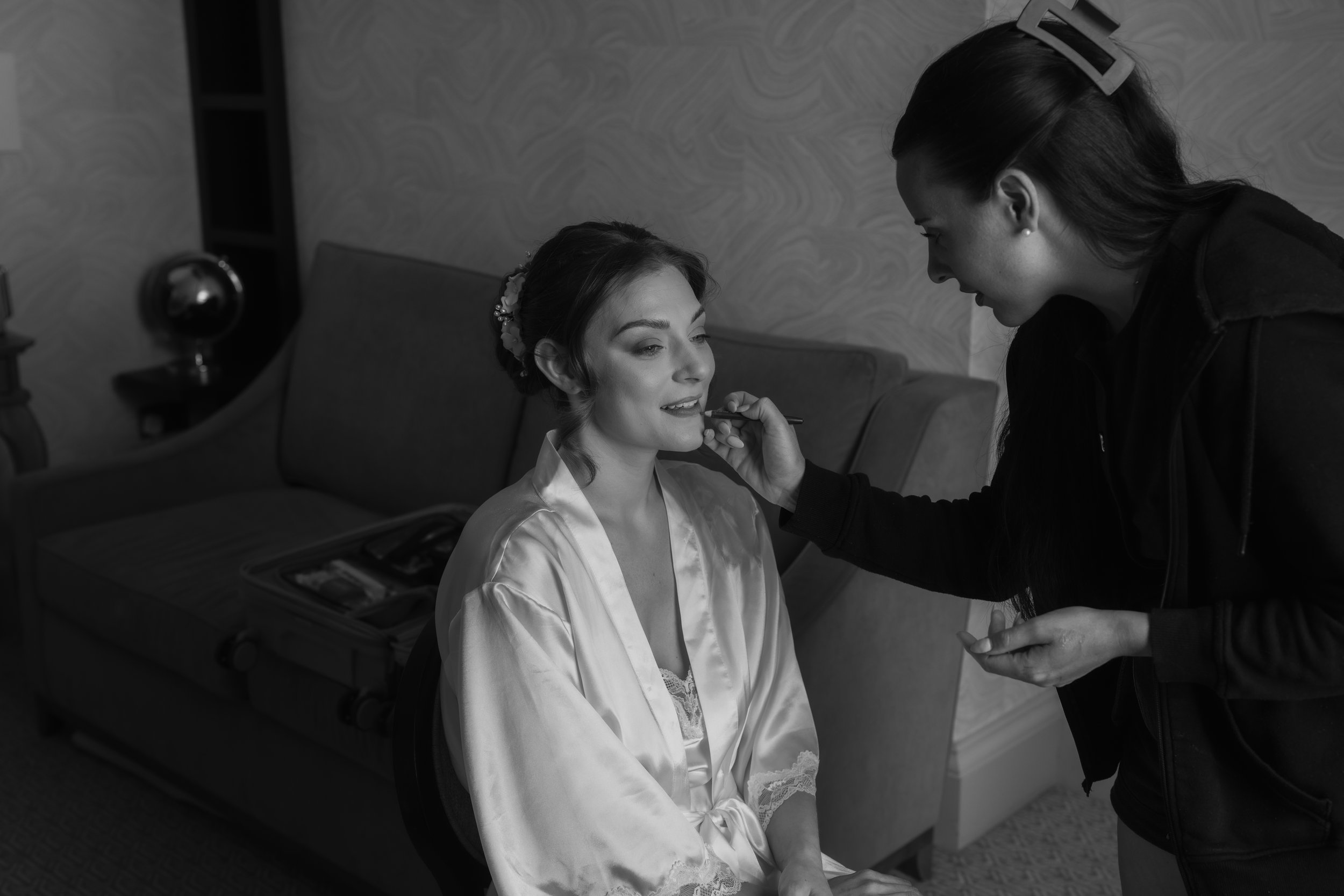A woman in a satin robe is having makeup applied by a makeup artist in a room with patterned wallpaper, a bench, and a globe on a side table.