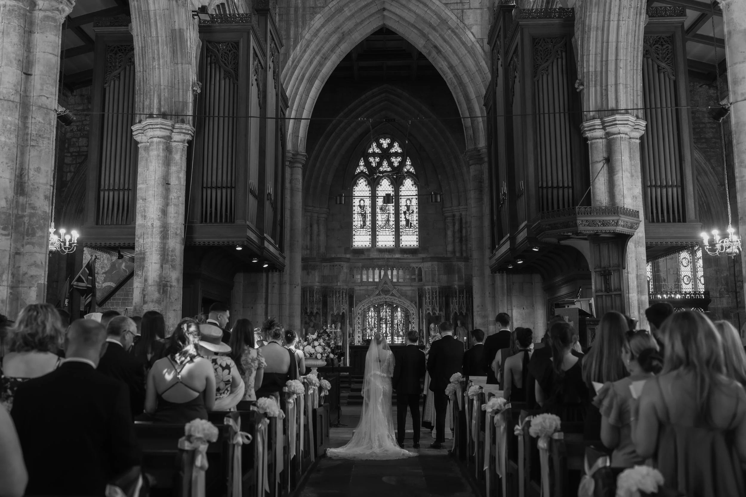 Interior of a church during a wedding ceremony with guests seated, bride and groom at the altar, stained glass window, and gothic architecture.